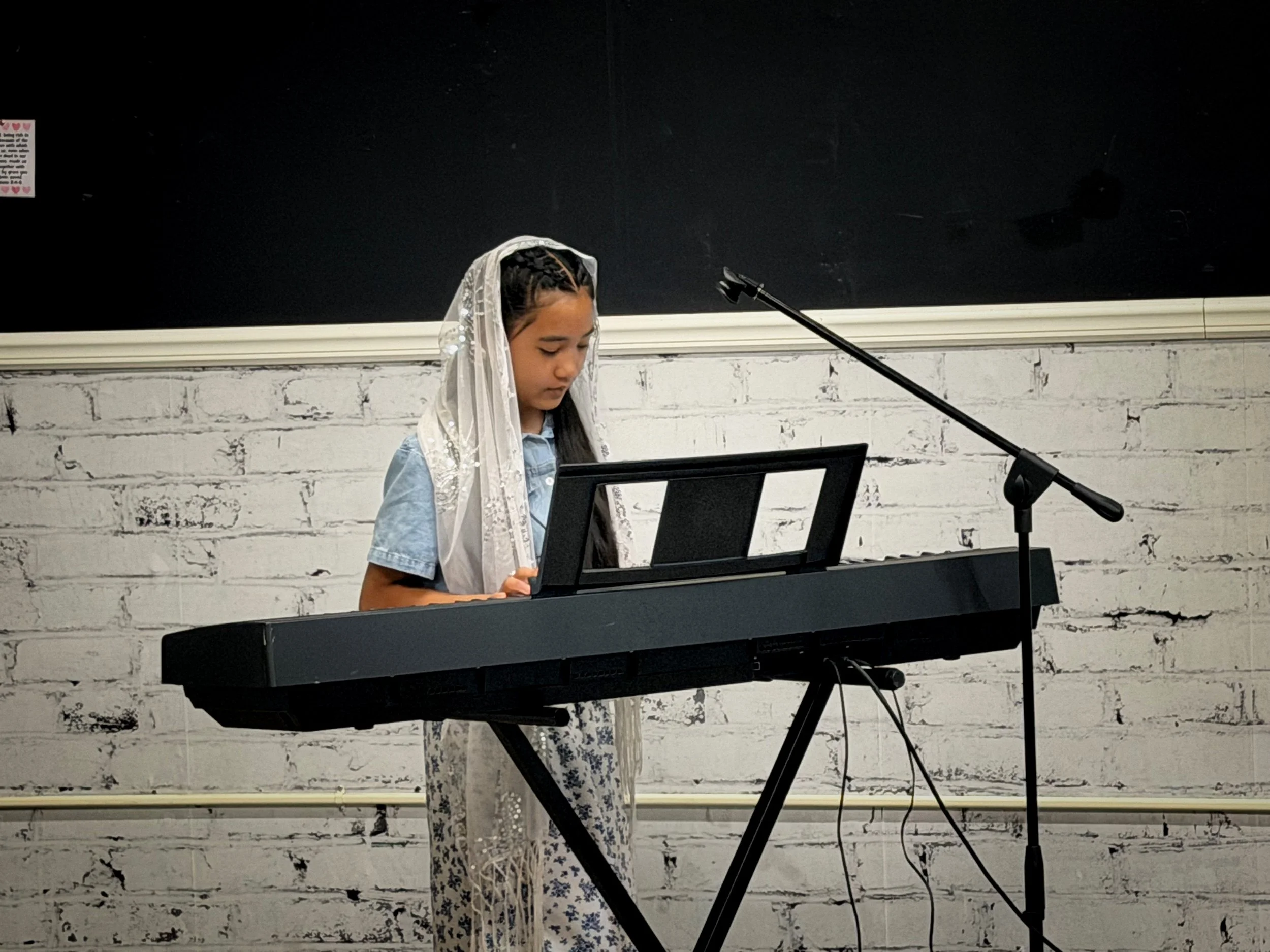 A young girl with braided hair, wearing a veil, playing a keyboard in front of a blackboard and a white brick wall.