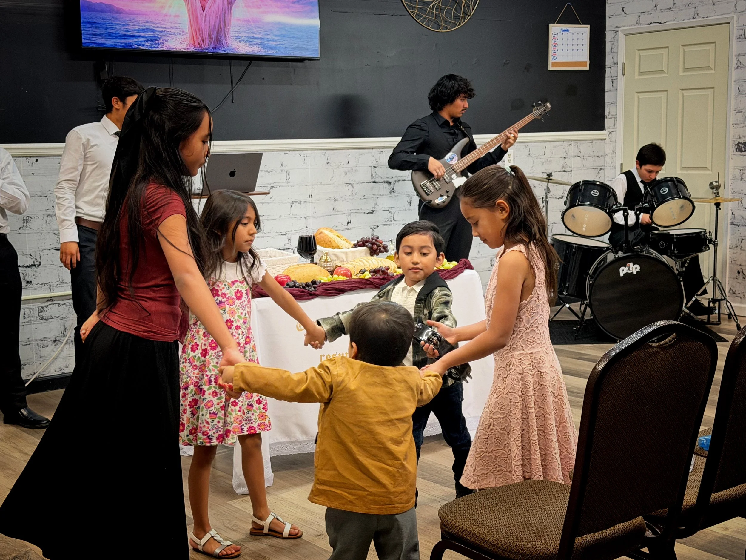 Children and young adults at a celebration or party with food and drinks, including grapes, bread, and a glass of wine, in a room with a brick wall and musical performers playing instruments.