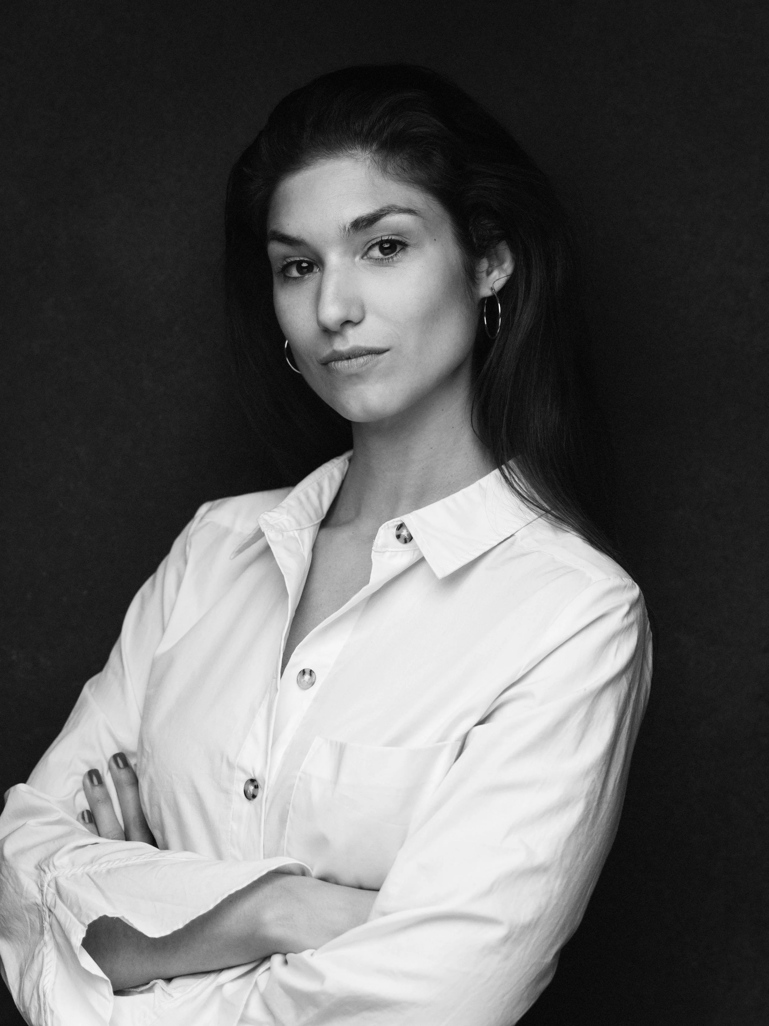 Black and white portrait of a woman with dark hair, wearing hoop earrings and a white shirt with rolled-up sleeves, standing against a dark background.