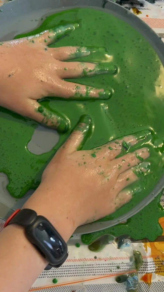Child's hands covered in green slime or paint in a plastic container.