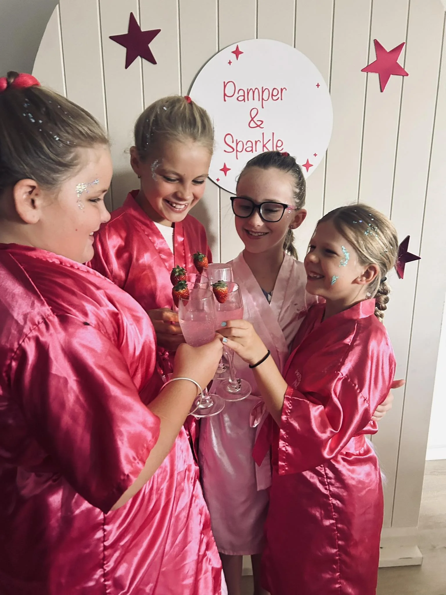 Four girls in pink satin robes celebrating with pink cocktails, some with strawberries, in front of a wall with a sign that says 'Pamper & Sparkle' and star decorations.