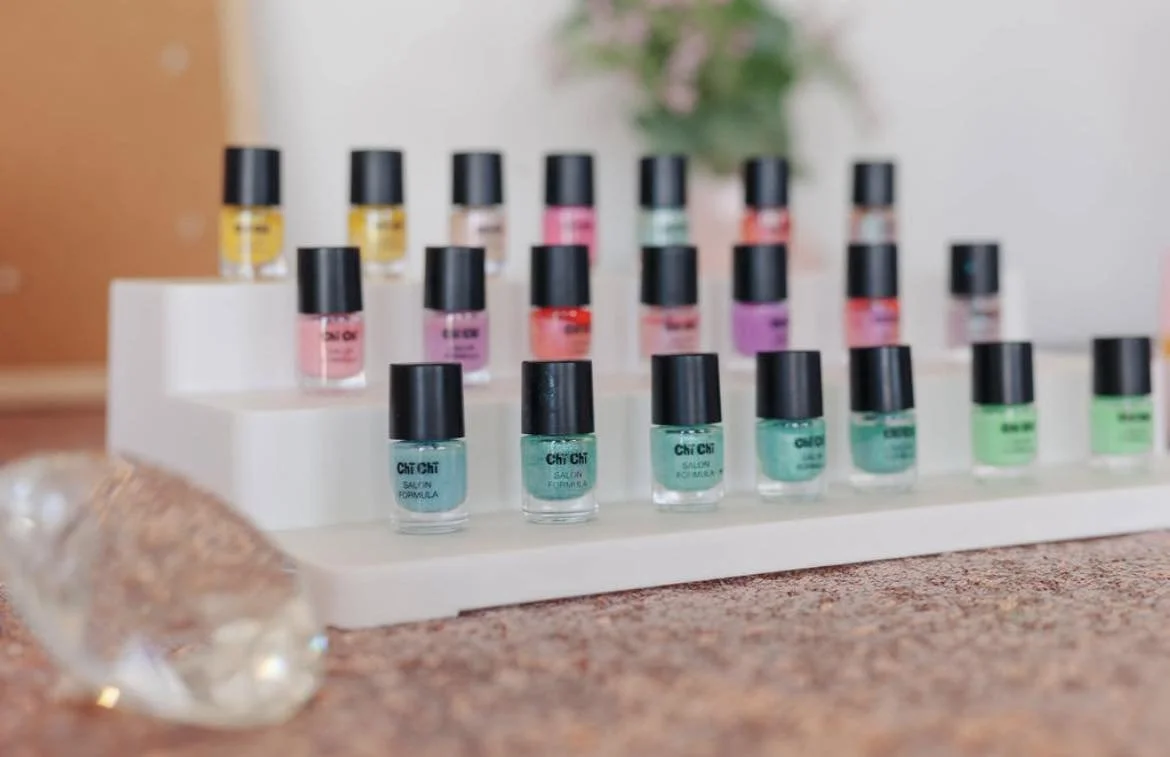 Assorted bottles of nail polish arranged on a white display stand, with a blurred plant in the background and a decorative glass object in the foreground.