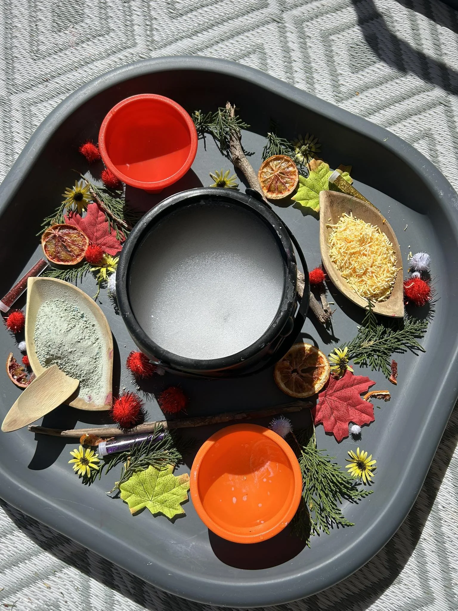 A circular tray containing various autumn-themed items, including three small bowls in red, orange, and black, a wooden dish with shredded cheese, another with powdered seasoning, dried leaves, small branches, and decorative flowers.