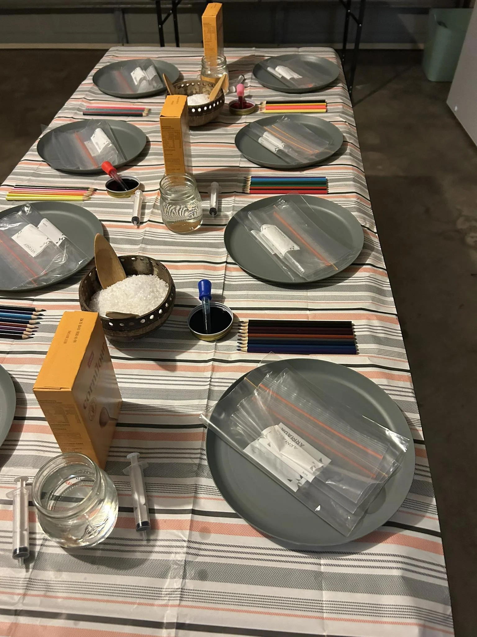 A dining table set with gray plates, plastic bags containing supplies, colored pencils, small bowls with black liquid, orange and yellow boxes, and various items for a science or art activity.