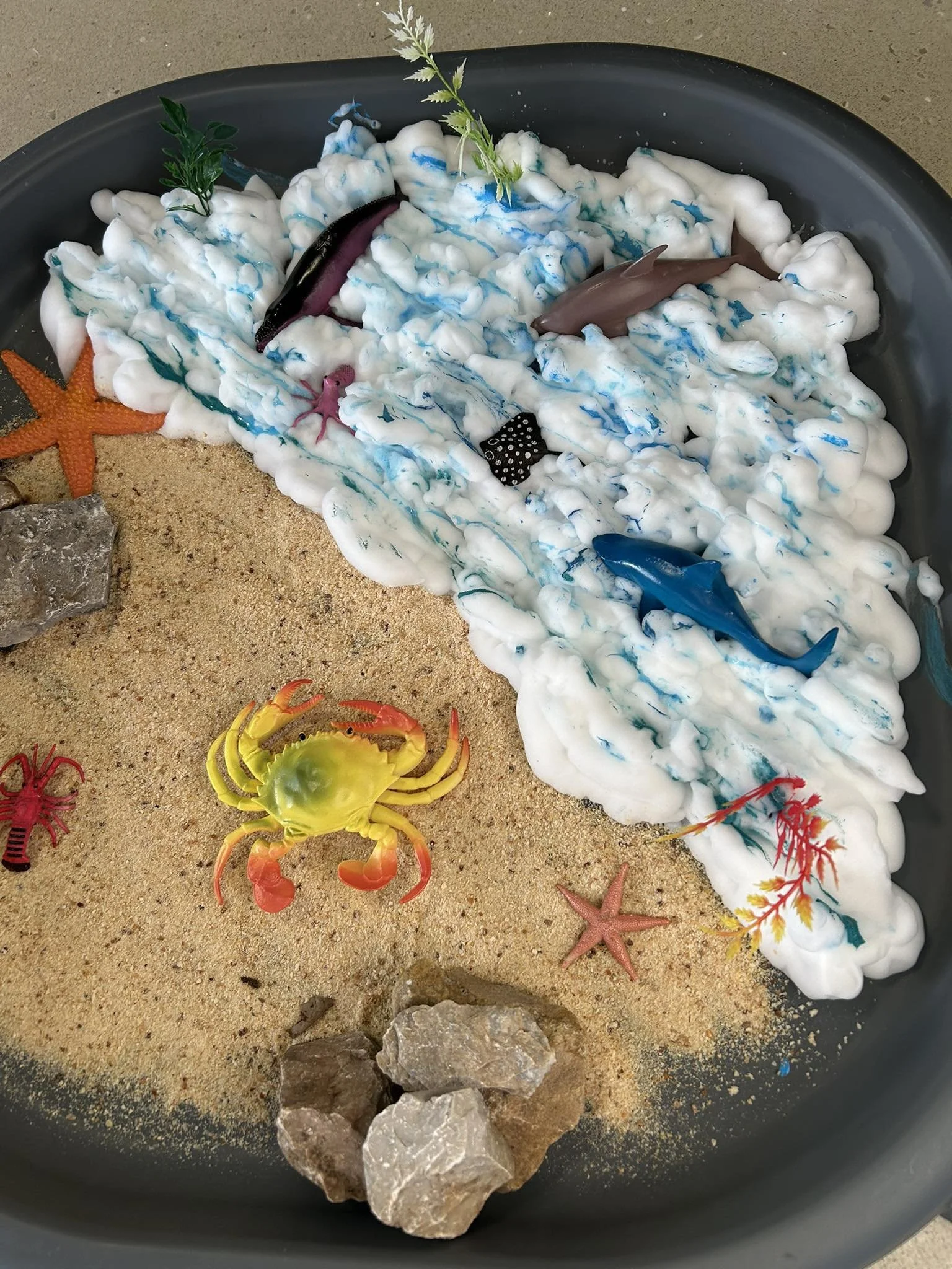 Decorative miniature ocean scene with toy dolphins, sharks, starfish, crabs, rocks, and foam representing water on sandy surface.