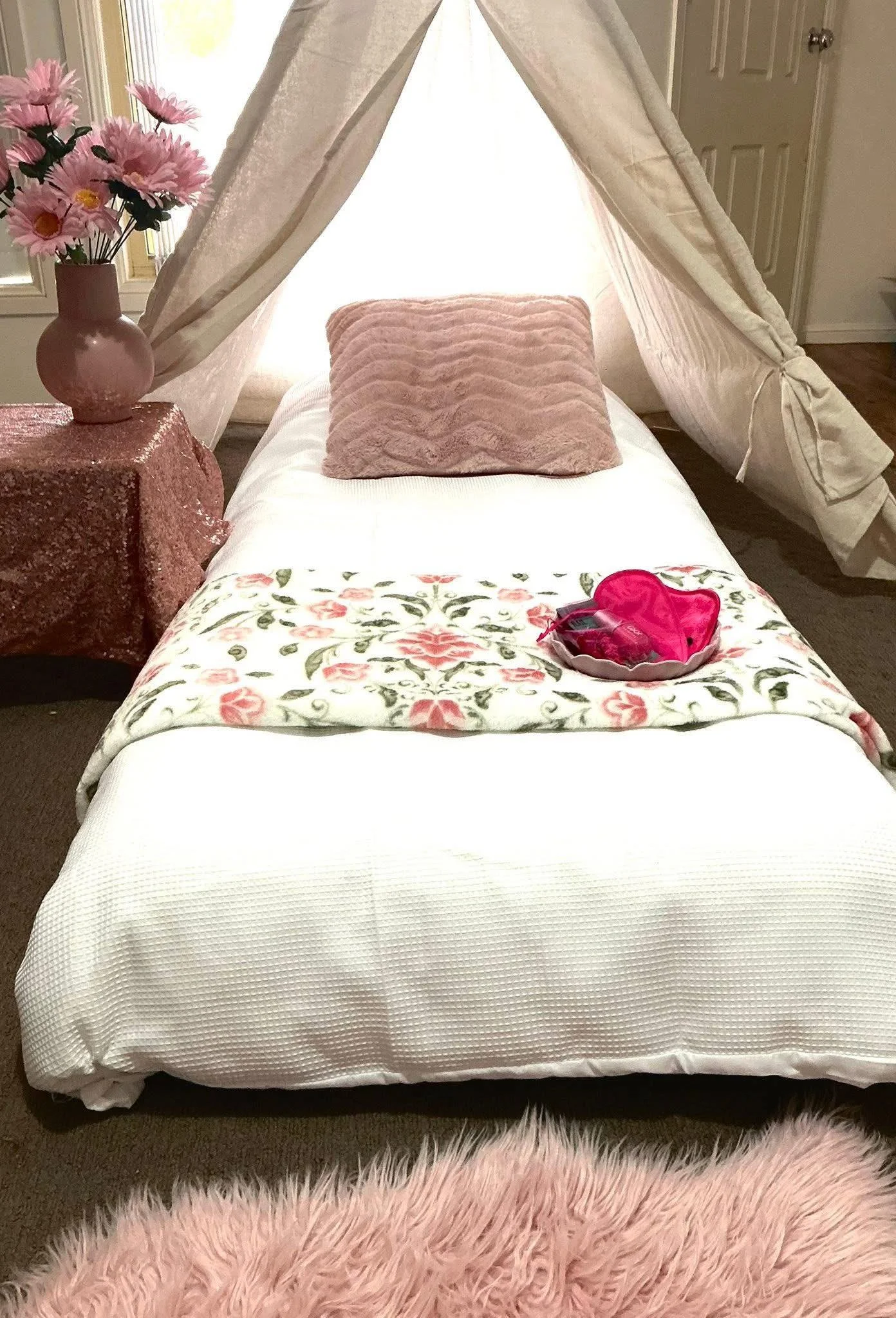 A cozy bedroom scene featuring a small bed with a white textured blanket, a pink velvet pillow, and a decorative blanket with floral print. There is a teepee-style canopy made of cream-colored fabric draped over the bed. A side table with pink sequined fabric holds a pink vase with pink artificial flowers. A pink heart-shaped box with a pair of pink slippers inside is placed on the bed. The room has a window with a view of trees outside and a door in the background.
