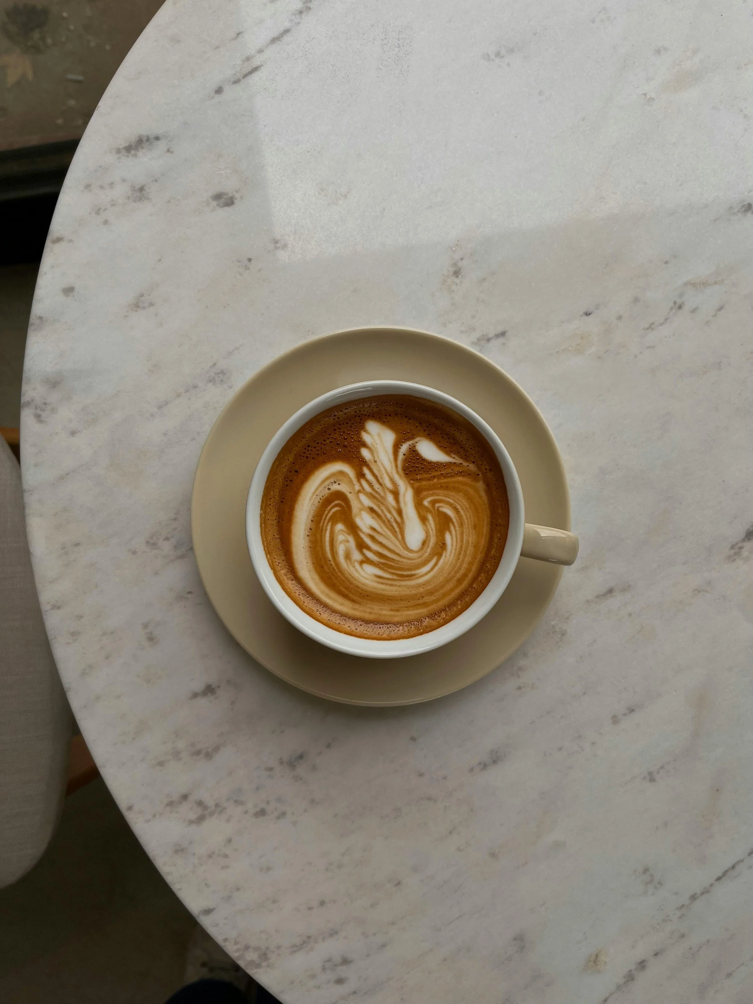A cup of coffee with latte art on top placed on a saucer on a white marble table.