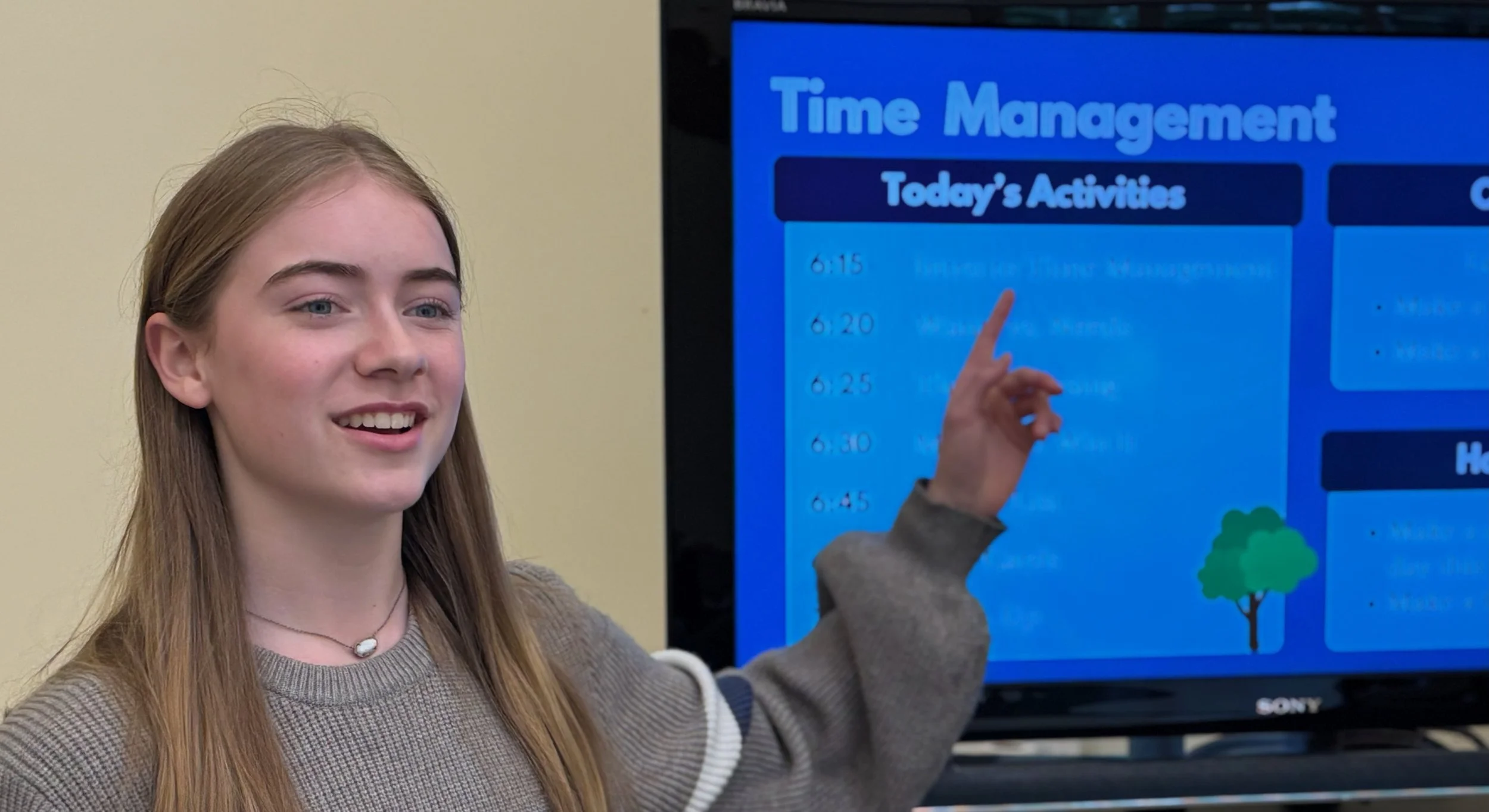 Teen Exec founder Natalya Lucas smiling, pointing at a digital display screen with a blue background, showing a schedule labeled 'Time Management' and 'Today's Activities'