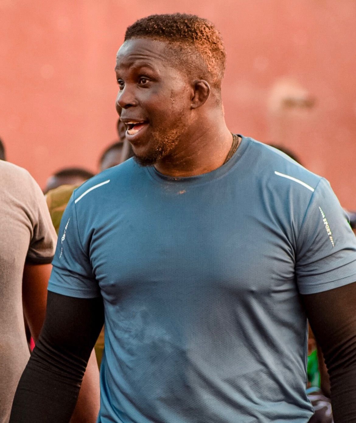 Pape Mbaye souriant portant un jersey bleu, entouré d'autres personnes, en plein air.