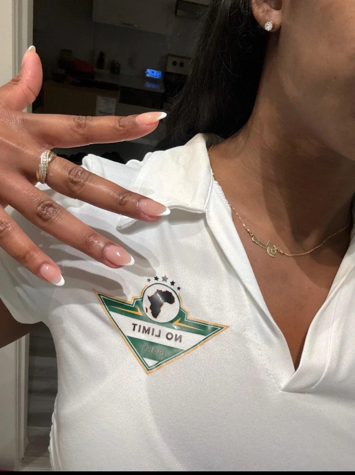 Une femme porte un collier avec un pendentif en forme de lettre 'S', une paire de boucles d'oreilles en diamants, une robe ou polo blanc avec un logo de la Ligue des Nations de football représentant l'Afrique, et montre ses mains avec des ongles en F