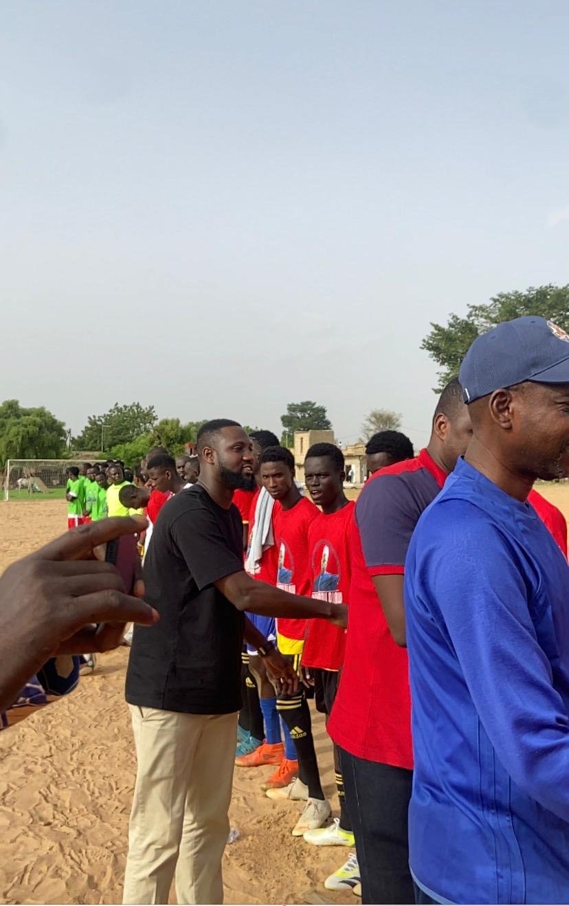 A man in a black t-shirt interacting with a line of young athletes on a sandy sports field; the athletes are wearing red and green jerseys during a community sports event.