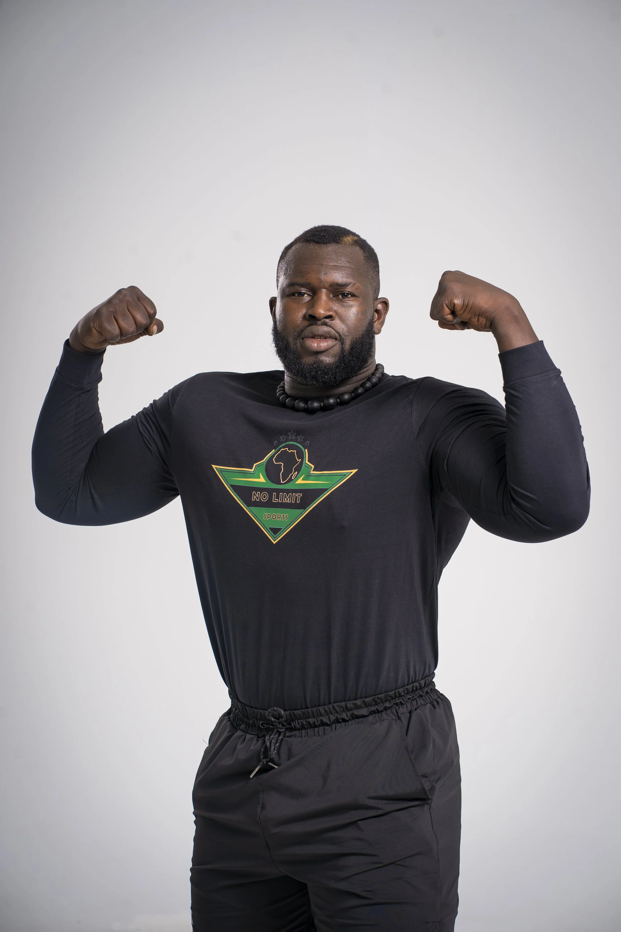 A powerful close-up of a professional athlete in a fighting stance, wearing a black long-sleeve No Limit Sports shirt featuring the green and gold shield logo with the African continent.