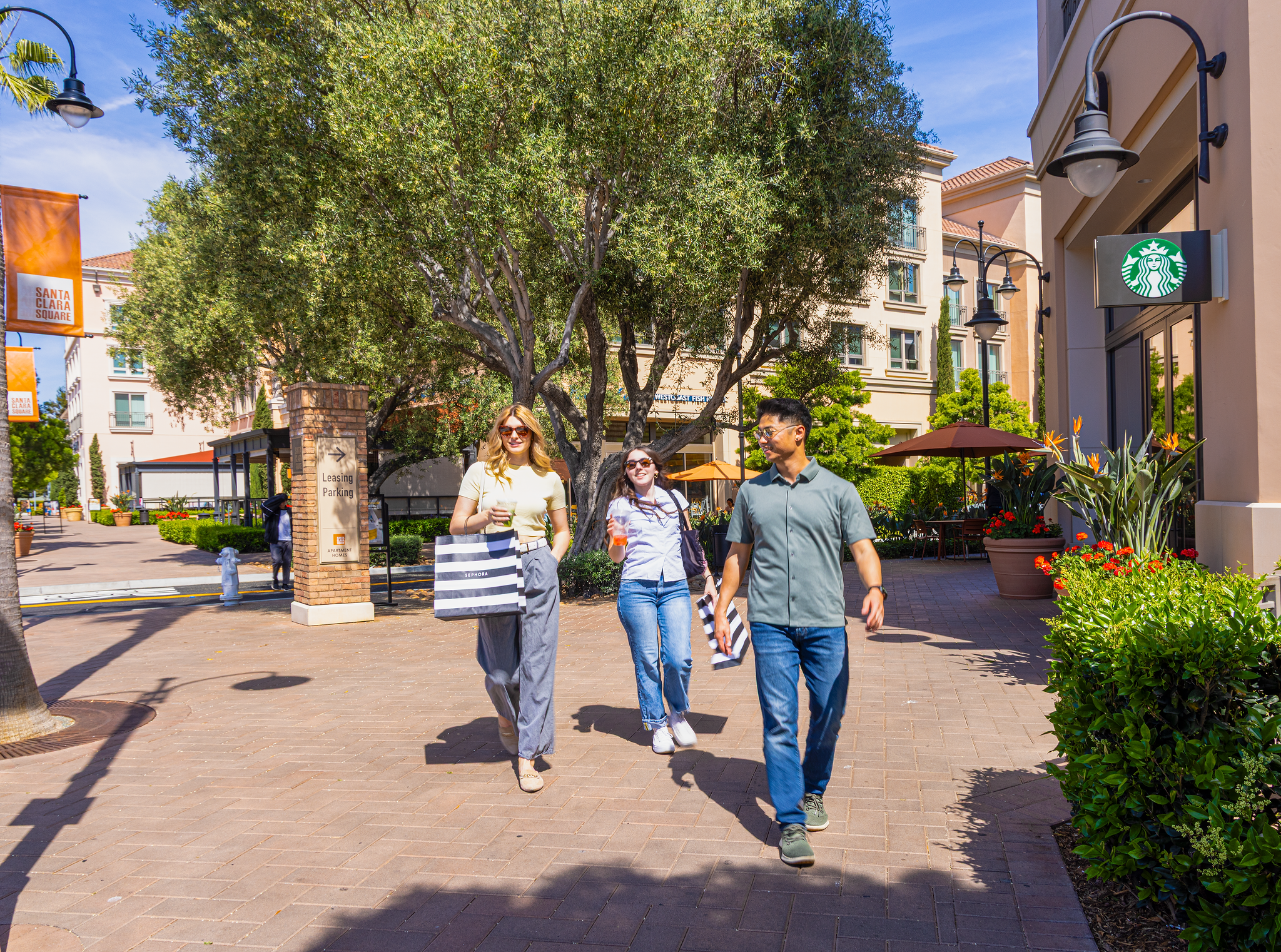 Four people walking outdoors in a shopping district with trees, shops, and outdoor seating. Two women and two men are smiling and enjoying a sunny day.
