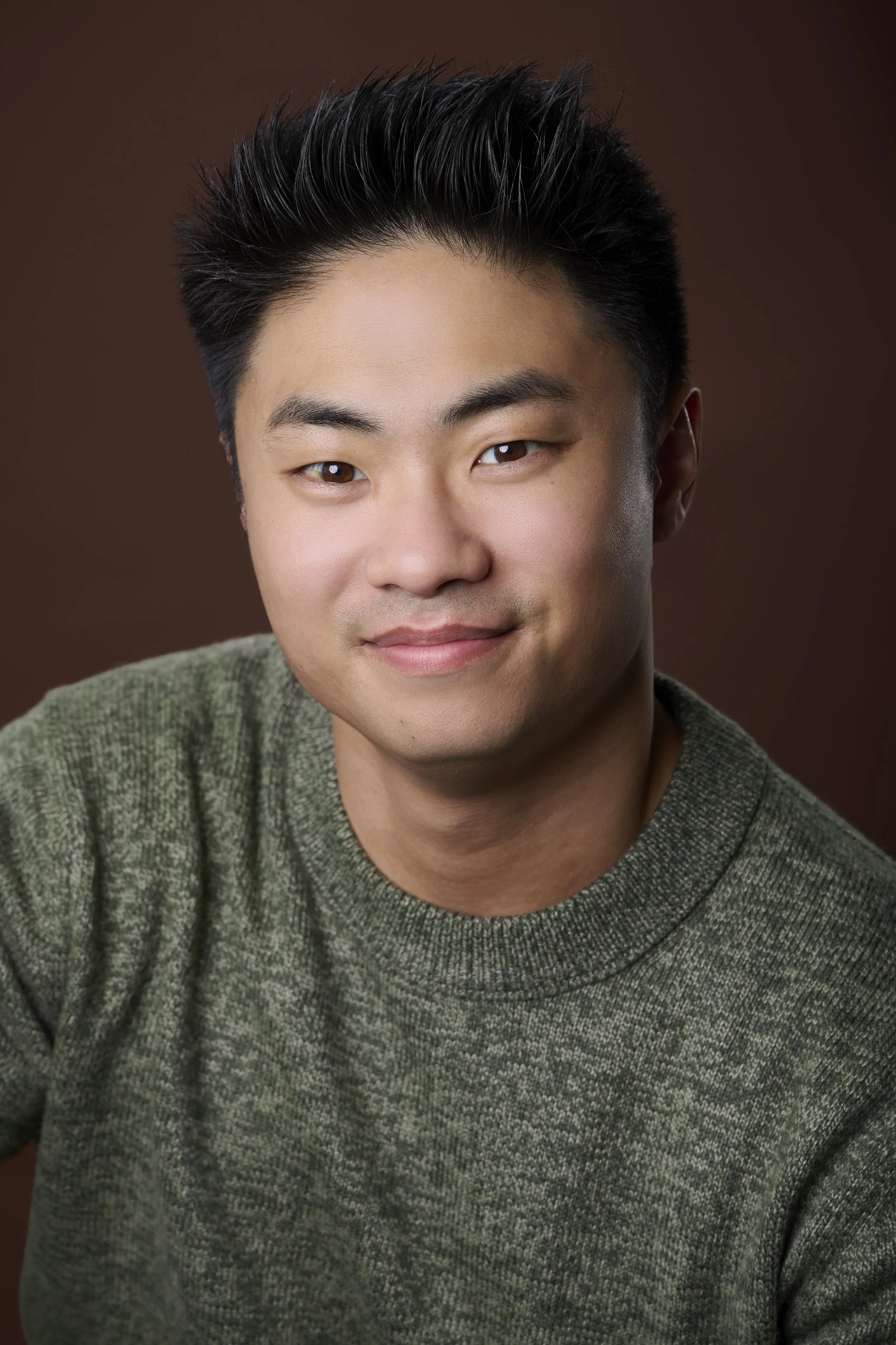 Close-up portrait of a young Asian man with short black hair, smiling, wearing a green textured sweater, against a brown background.