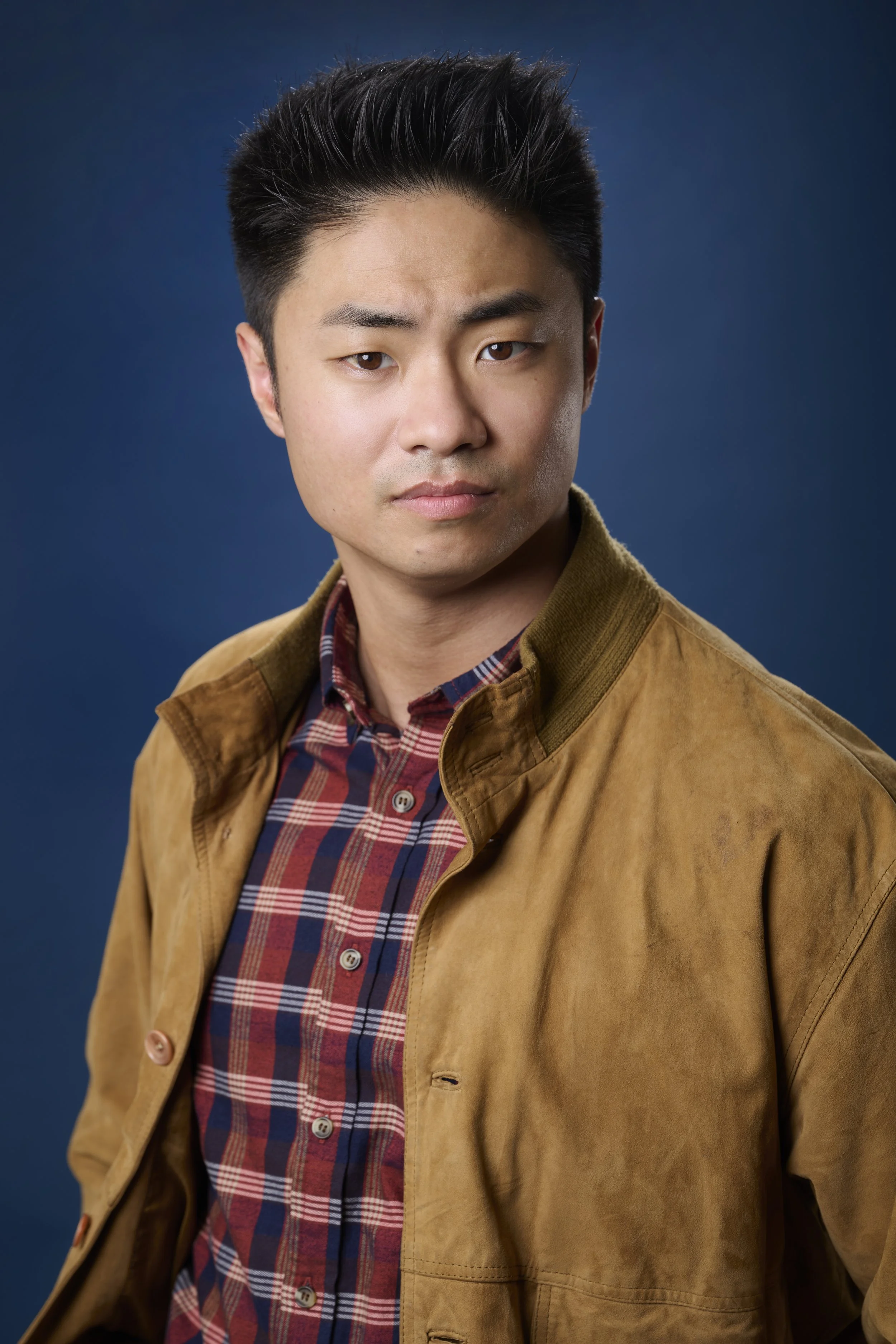 A young man with short black hair, wearing a tan jacket over a red, blue, and white plaid shirt, posing against a dark blue background.