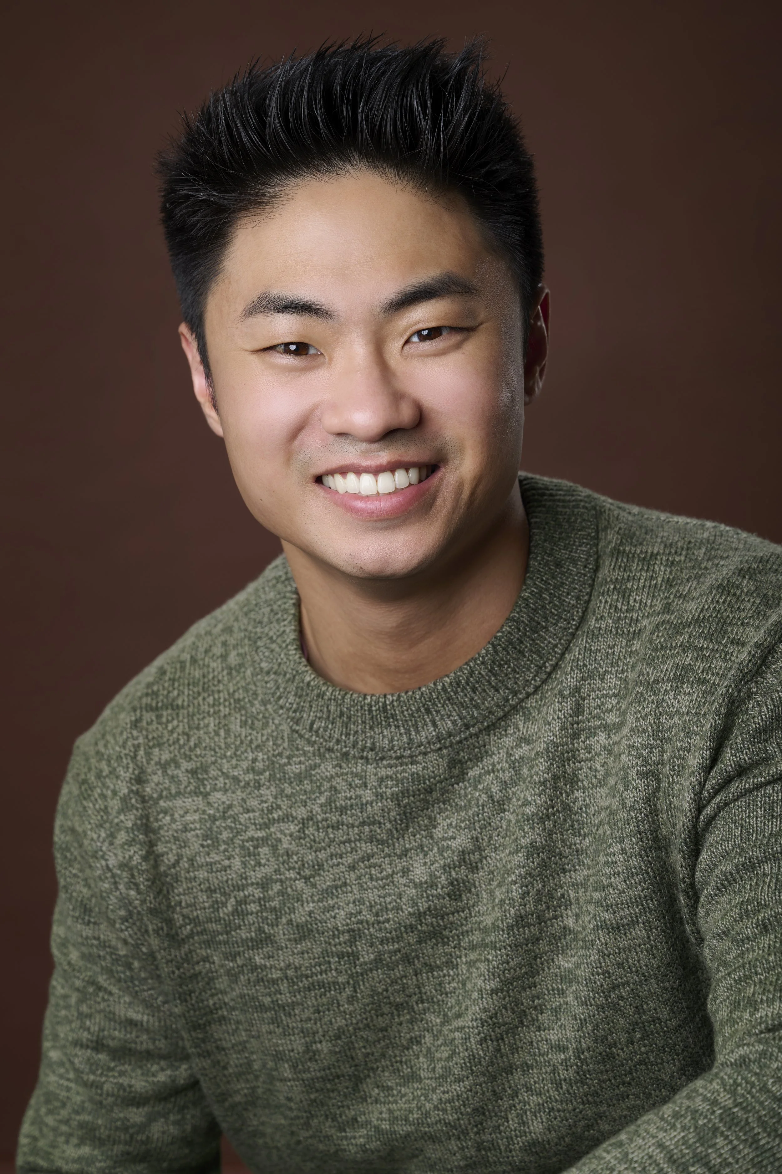 Headshot of a young Asian man with short, dark hair, smiling, wearing a green knit sweater, against a brown background.