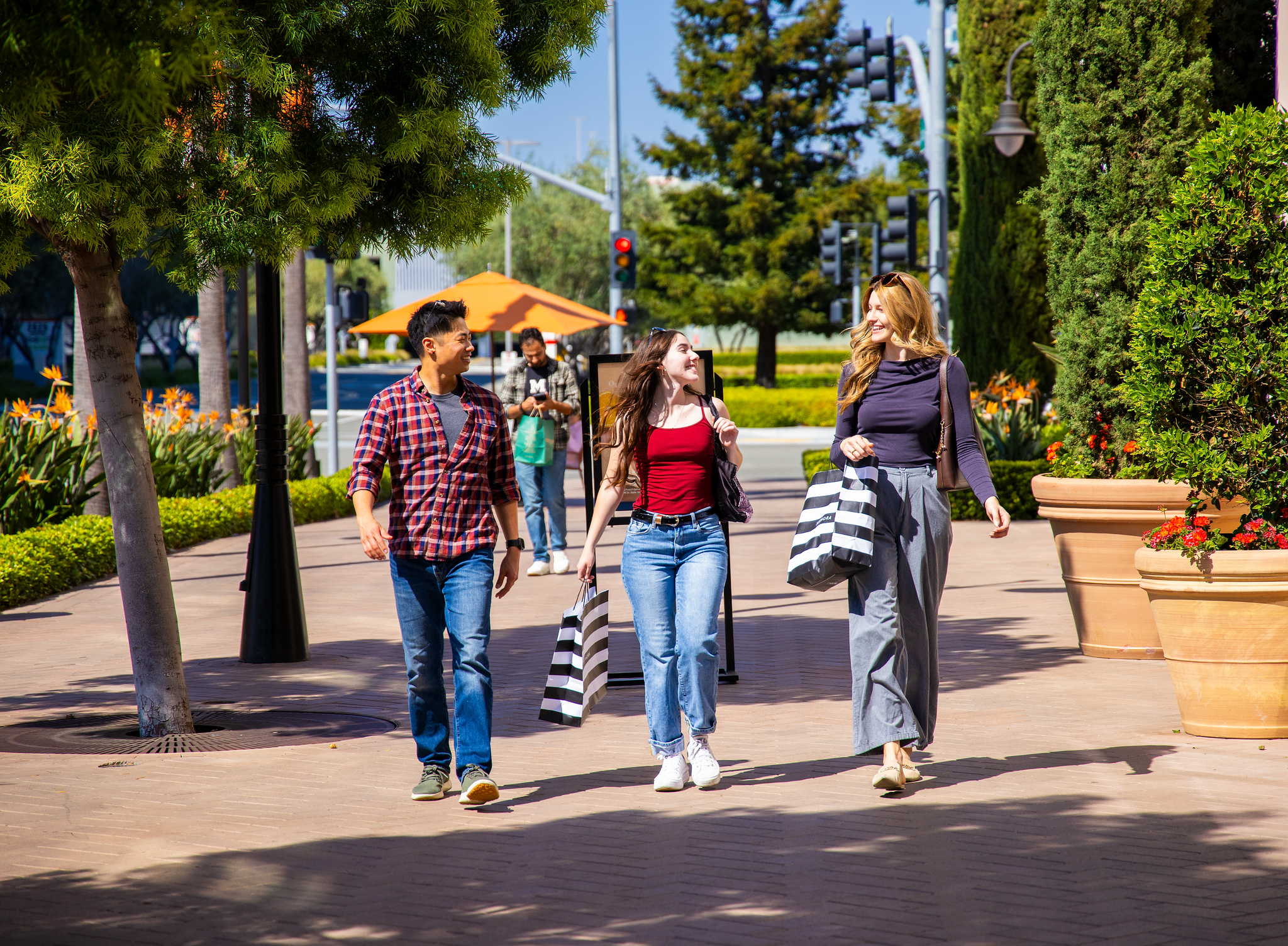 Four young people walking and talking outdoors in a sunny urban park or sidewalk area, surrounded by trees and large flower pots, with colorful umbrellas and streetlights in the background.