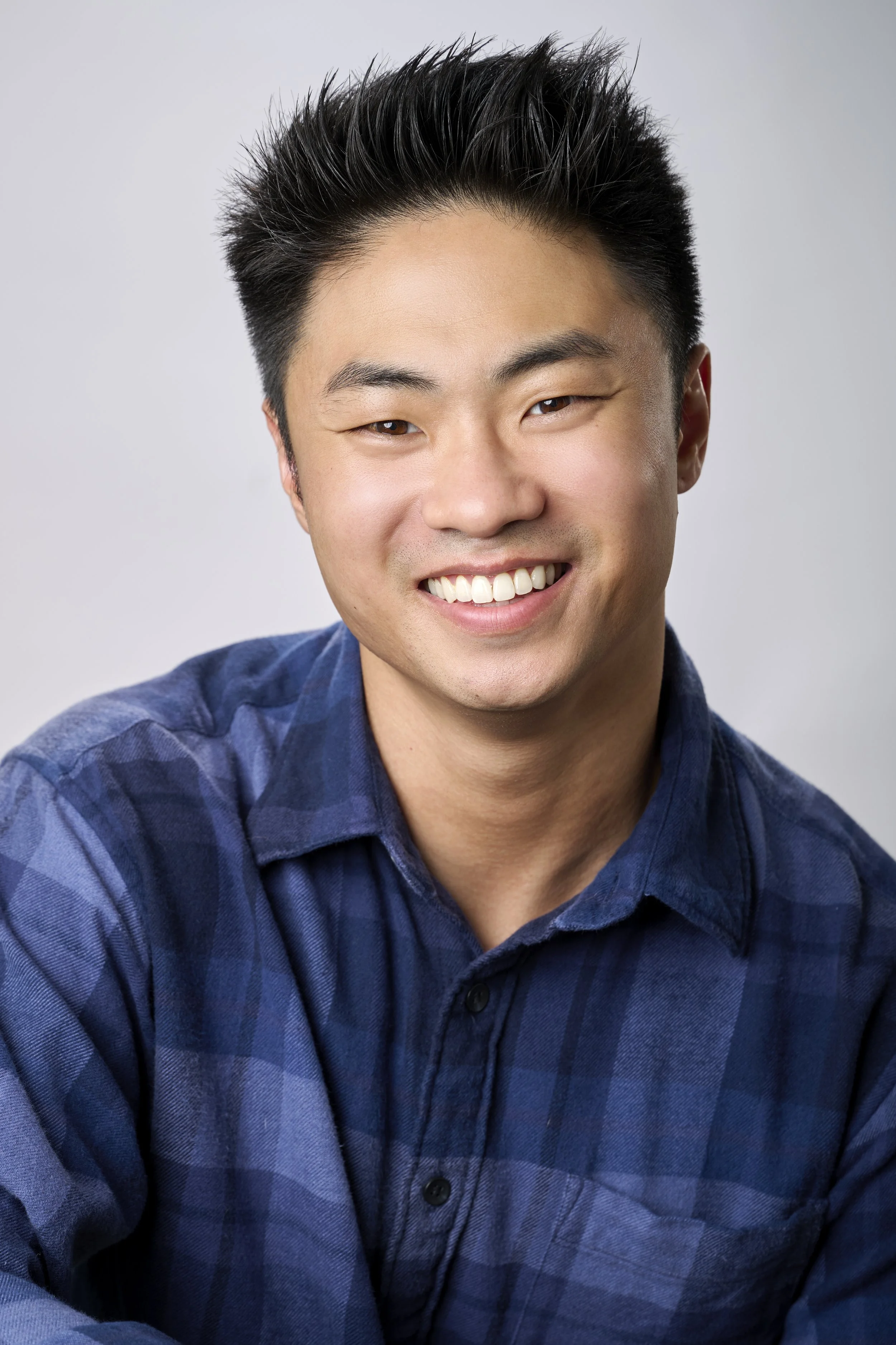 A young man with black spiked hair and a wide smile wearing a blue plaid shirt.