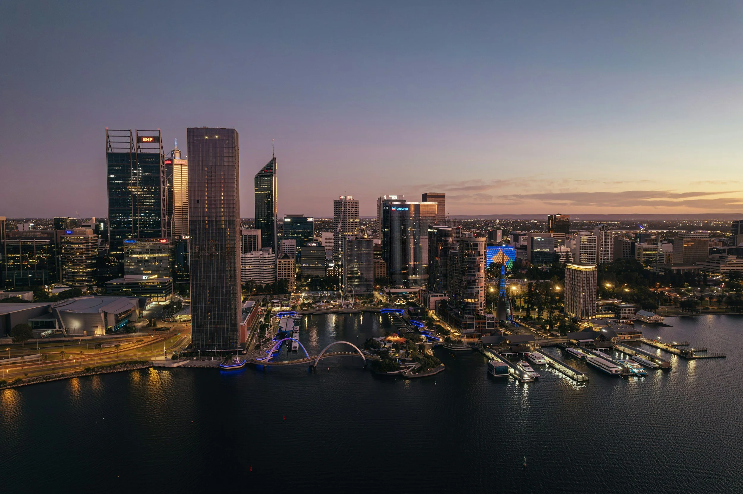 Downtown skyline of a city at sunset with tall skyscrapers, a marina filled with boats, and a waterfront promenade with illuminated walkways.