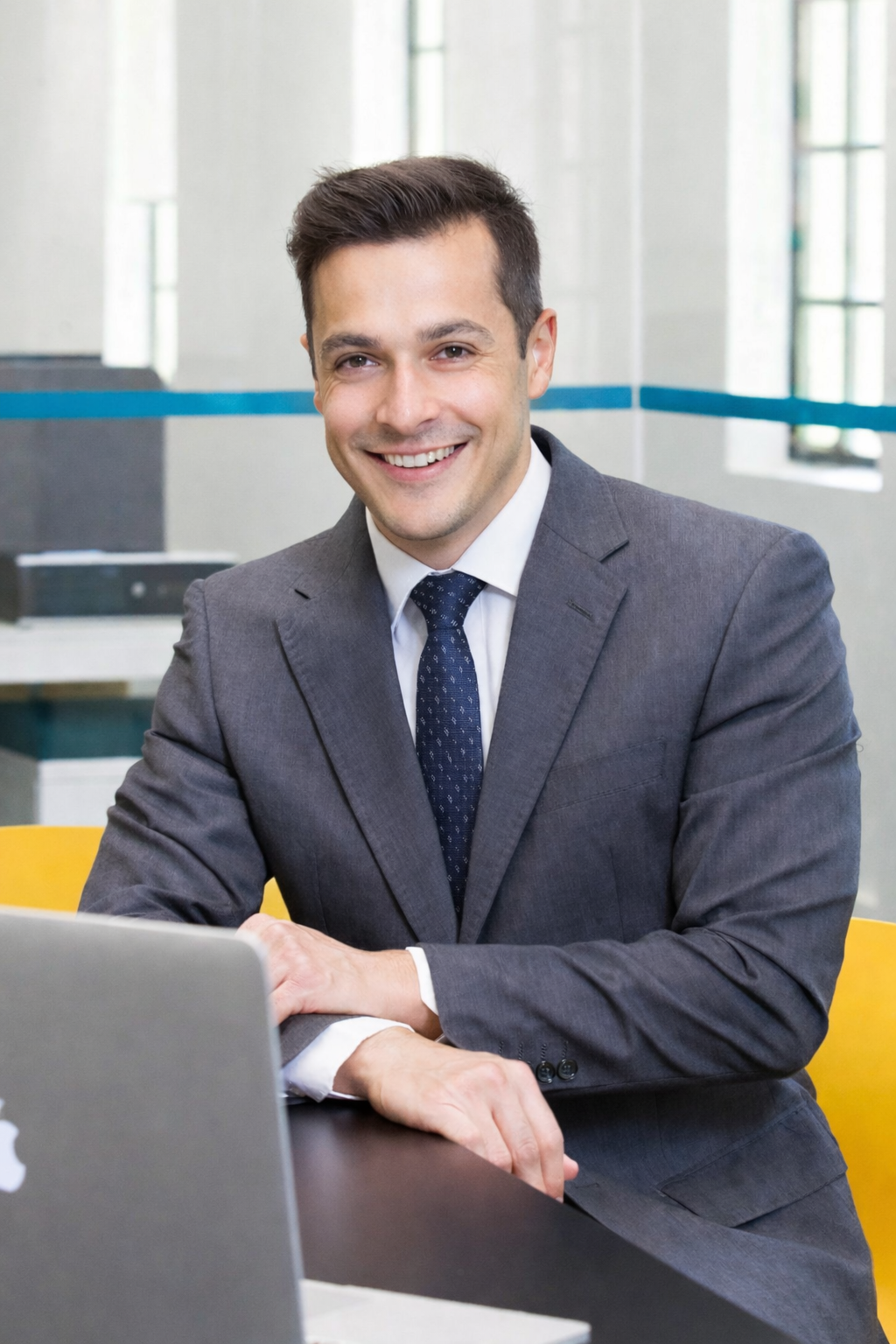 A smiling man in a gray business suit sitting at a desk with a laptop in an office setting.