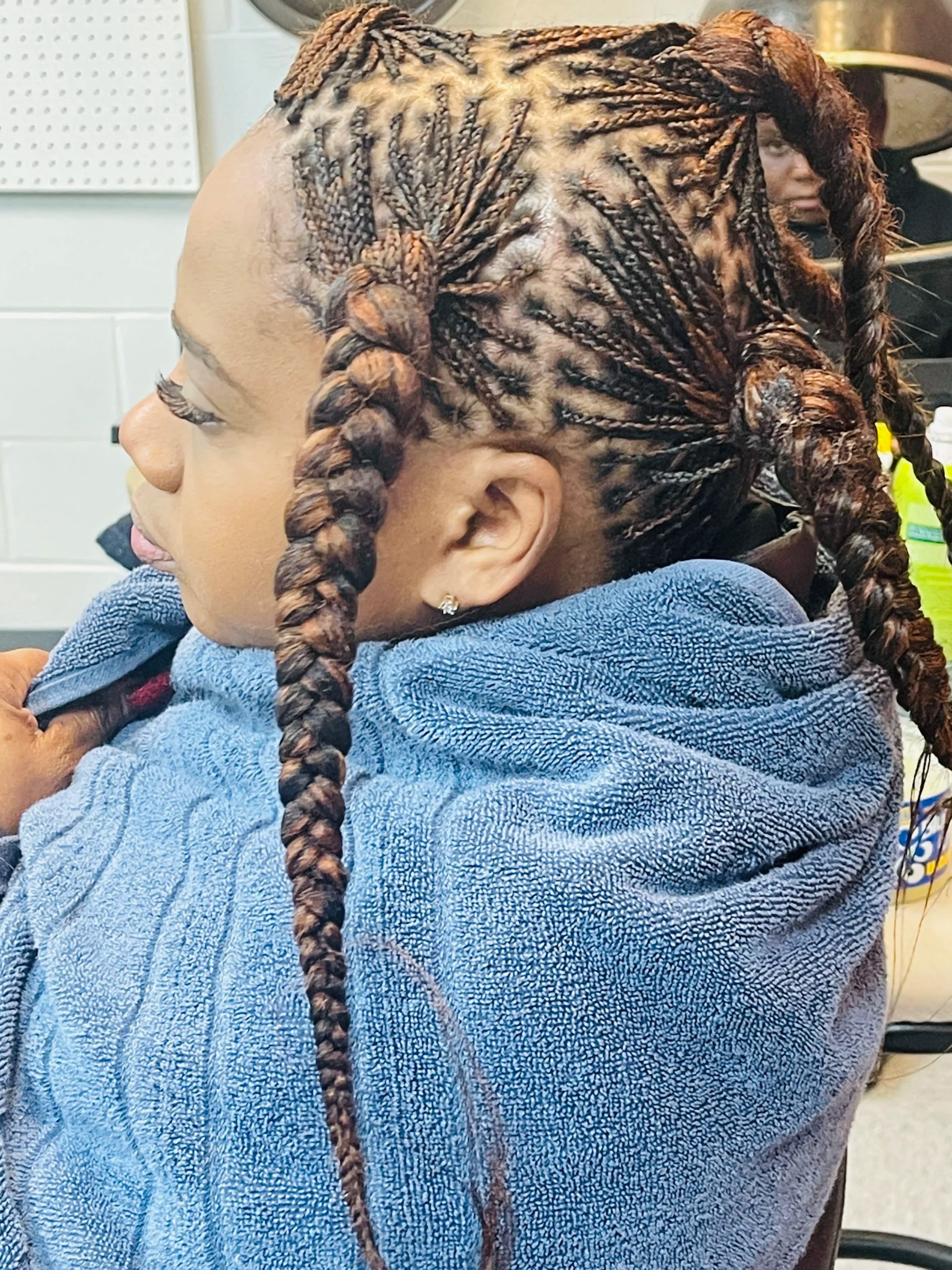 Side view of a woman with braided hair, sitting in a salon or barbershop, wrapped in a blue towel, with professional hair styling equipment in the background.