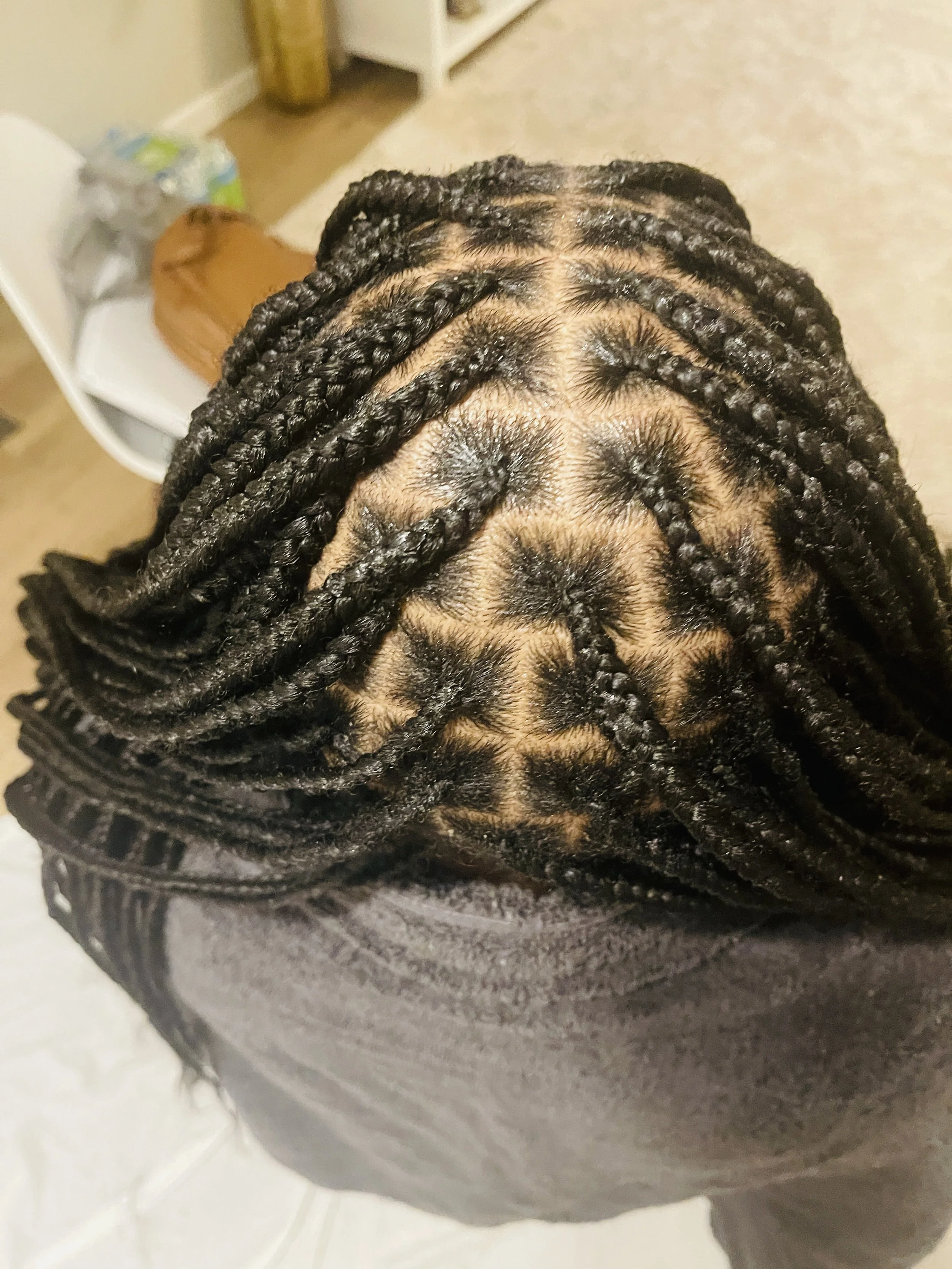 Top view of neatly styled black braided hair on a person's head with a visible parting in a room with a carpeted floor and furniture.