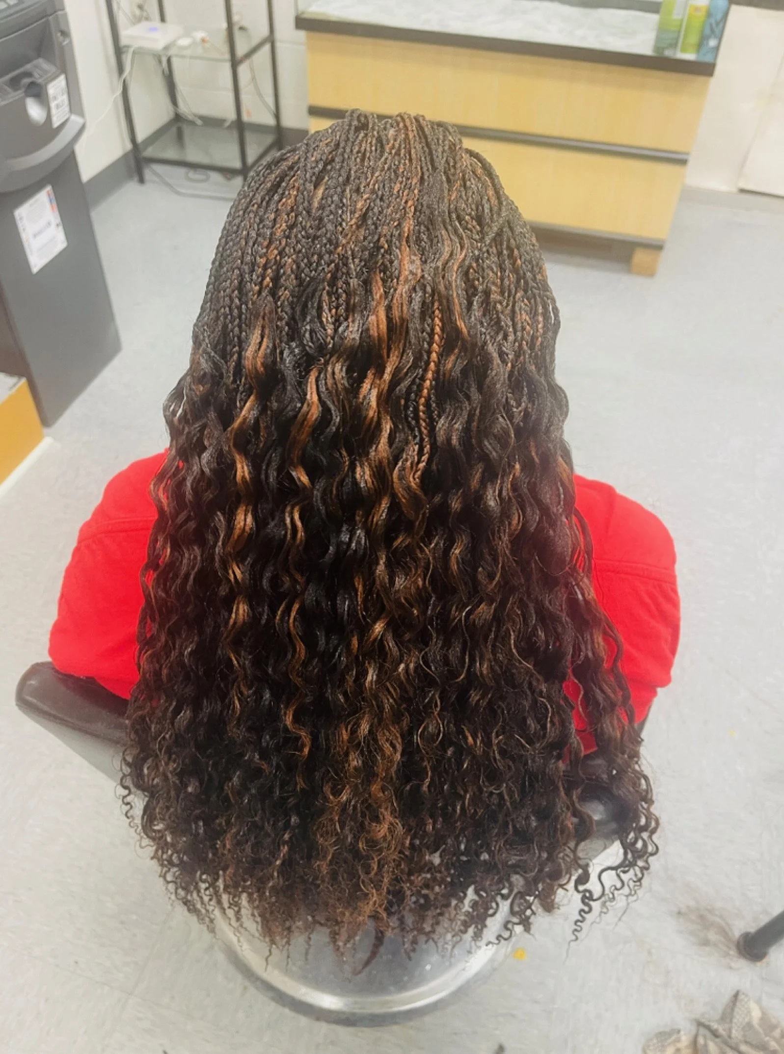 A person with long, styled braids and curly hair, sitting in a salon chair, wearing a red shirt, facing away from the camera in a salon or barbershop environment.