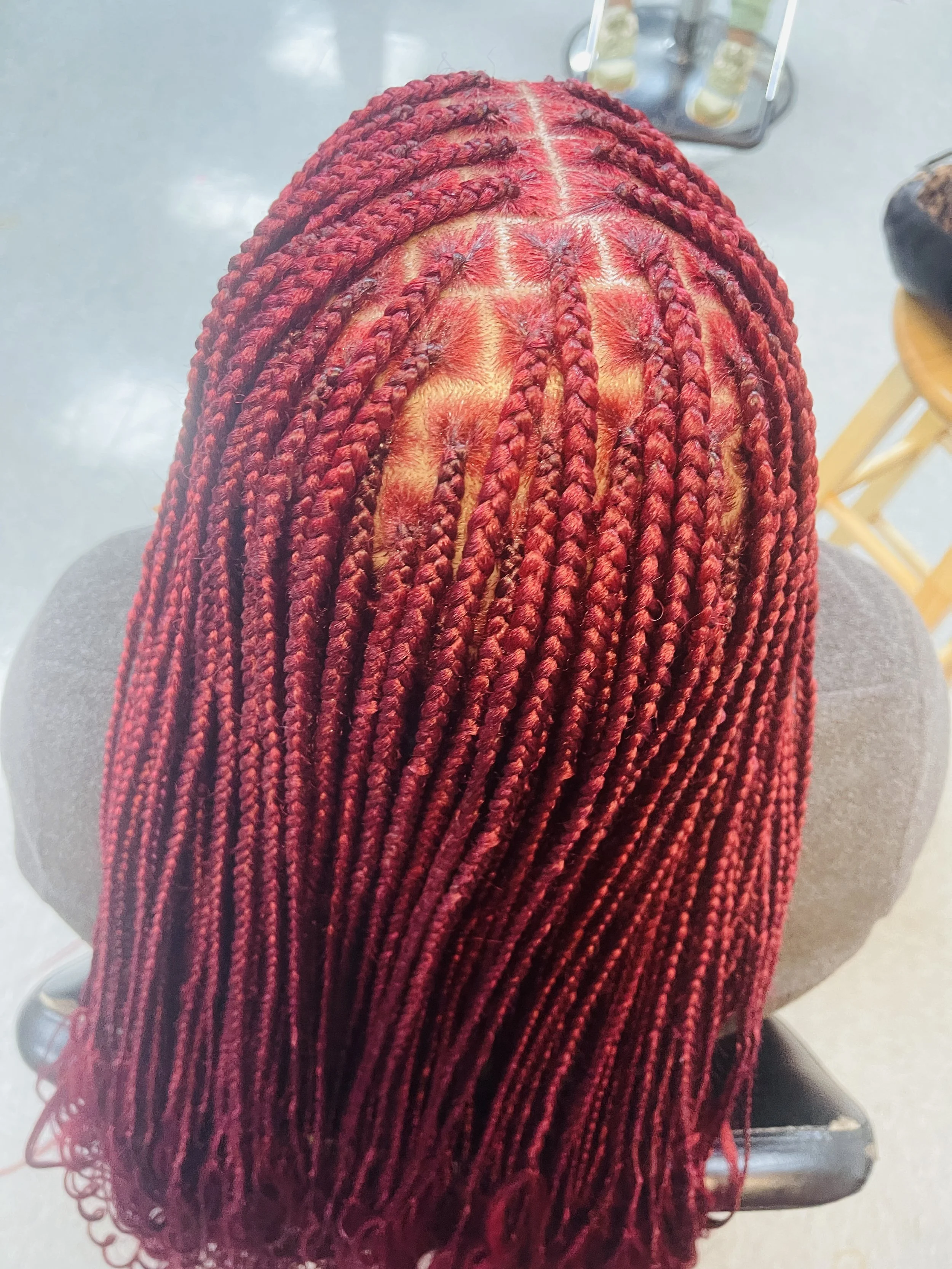 A top view of a person's head showing red braids with black beads at the ends. The background shows a gray floor, a chair, and some items on a tray.