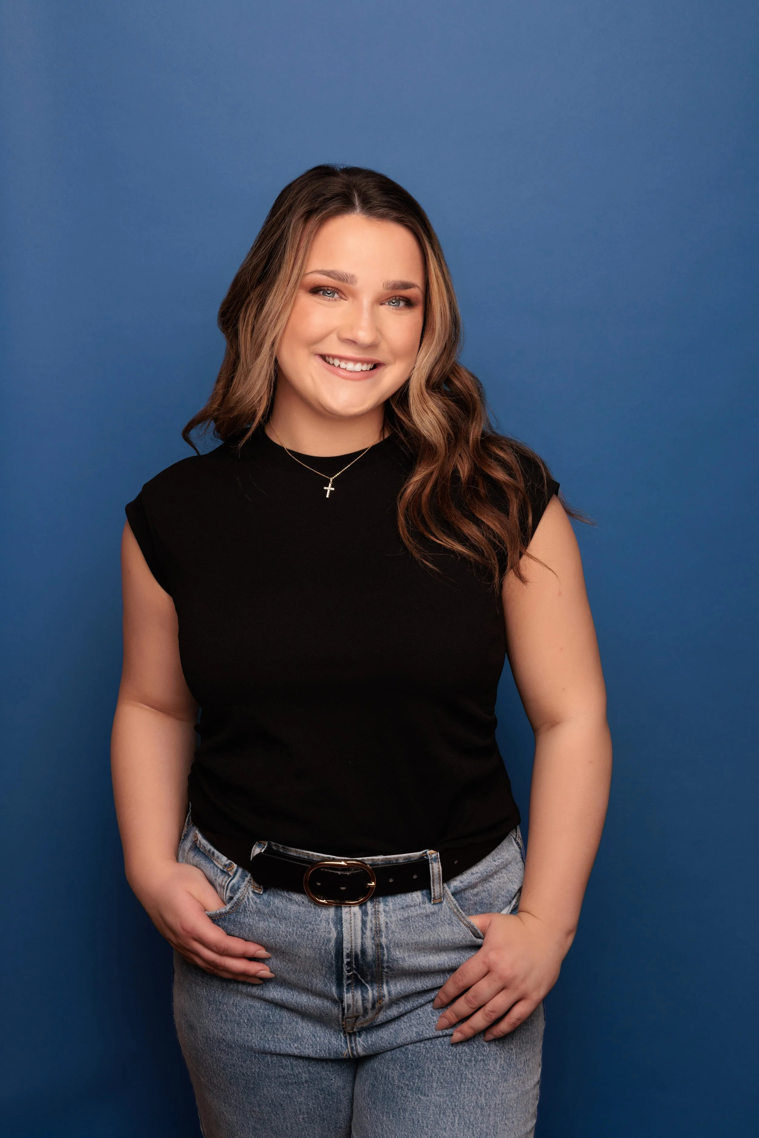 Young woman with wavy brown hair, wearing a black sleeveless top, blue jeans with a black belt, smiling, against a solid blue background.