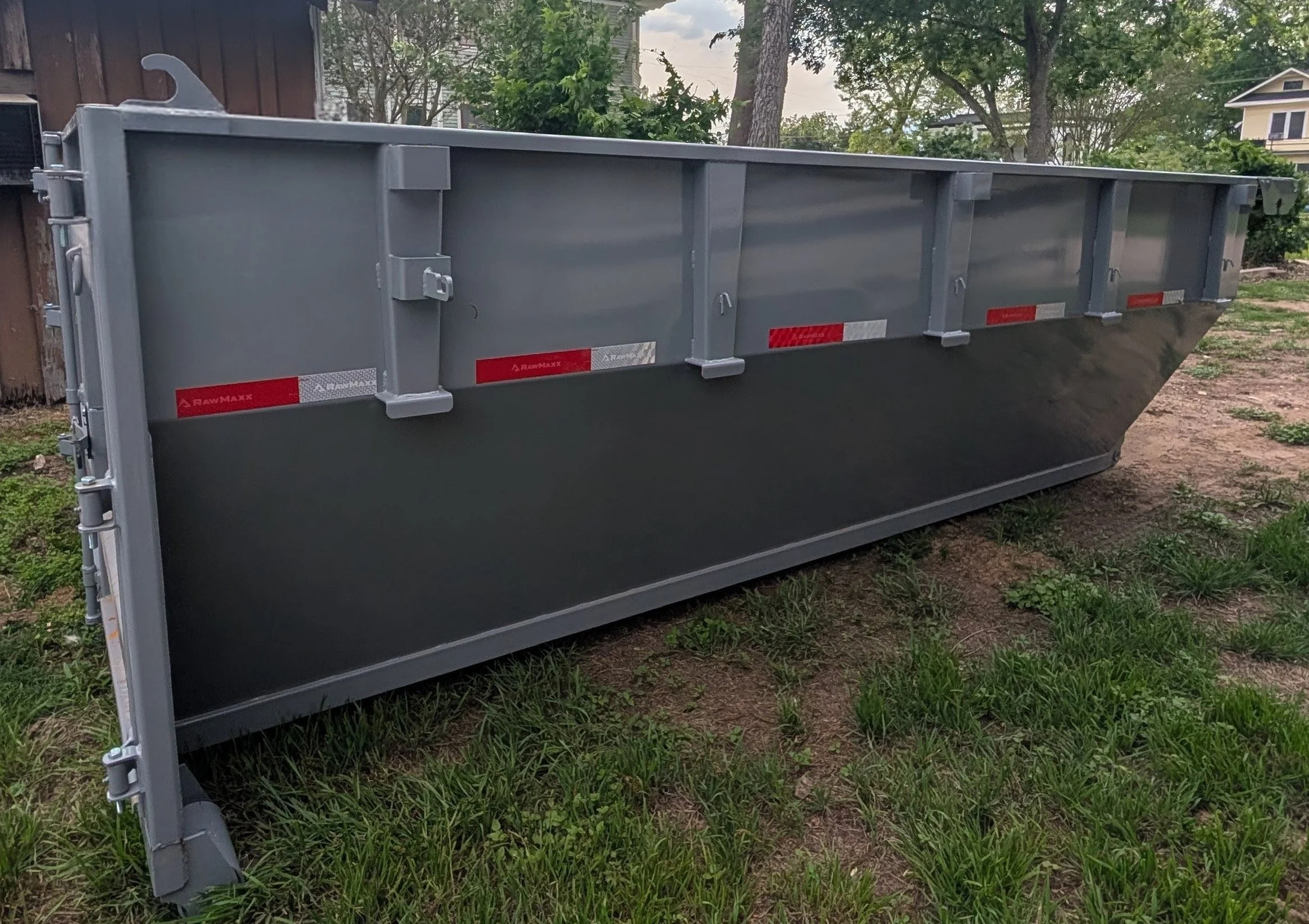 A large gray metal dumpster with red and white reflective labels, situated on a patch of grass with some dirt patches. In the background are trees and residential houses.