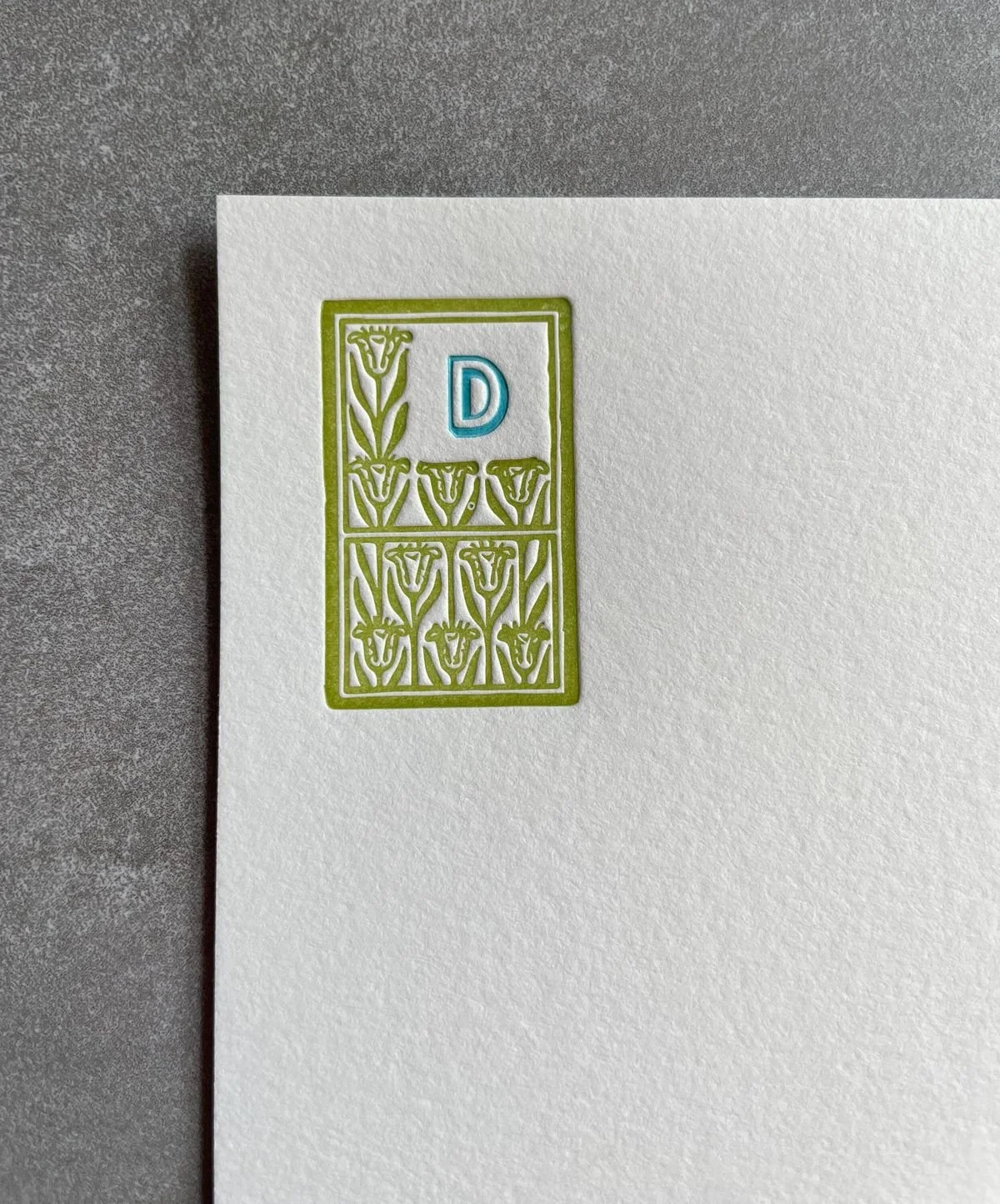 A white sheet of paper with an embossed green border and floral pattern, containing a blue letter 'D' in the upper right corner.