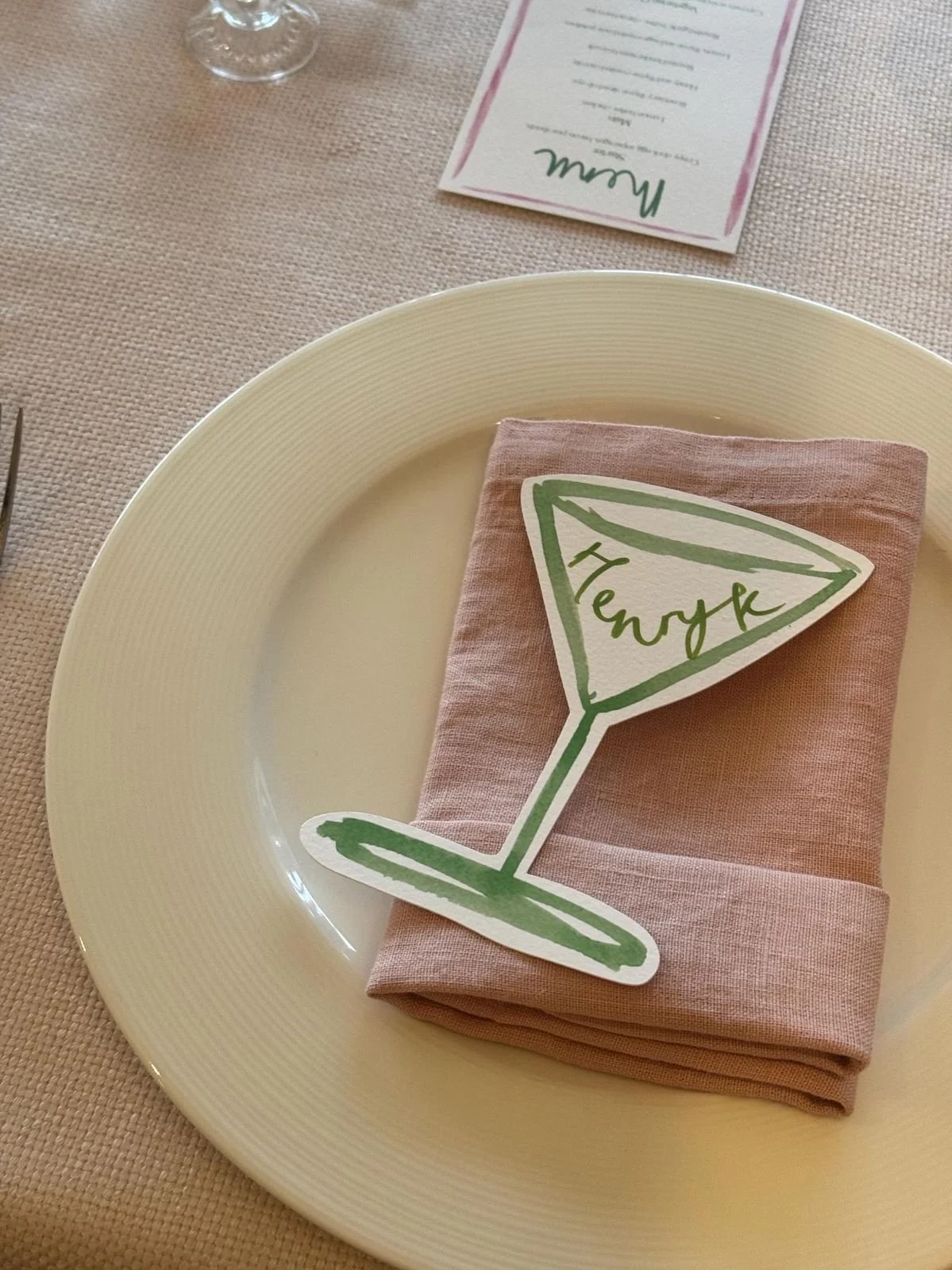 Place setting with a white plate, a pink napkin, and a paper cutout of a wine glass with the word "Henrik" written on it, on a beige tablecloth.