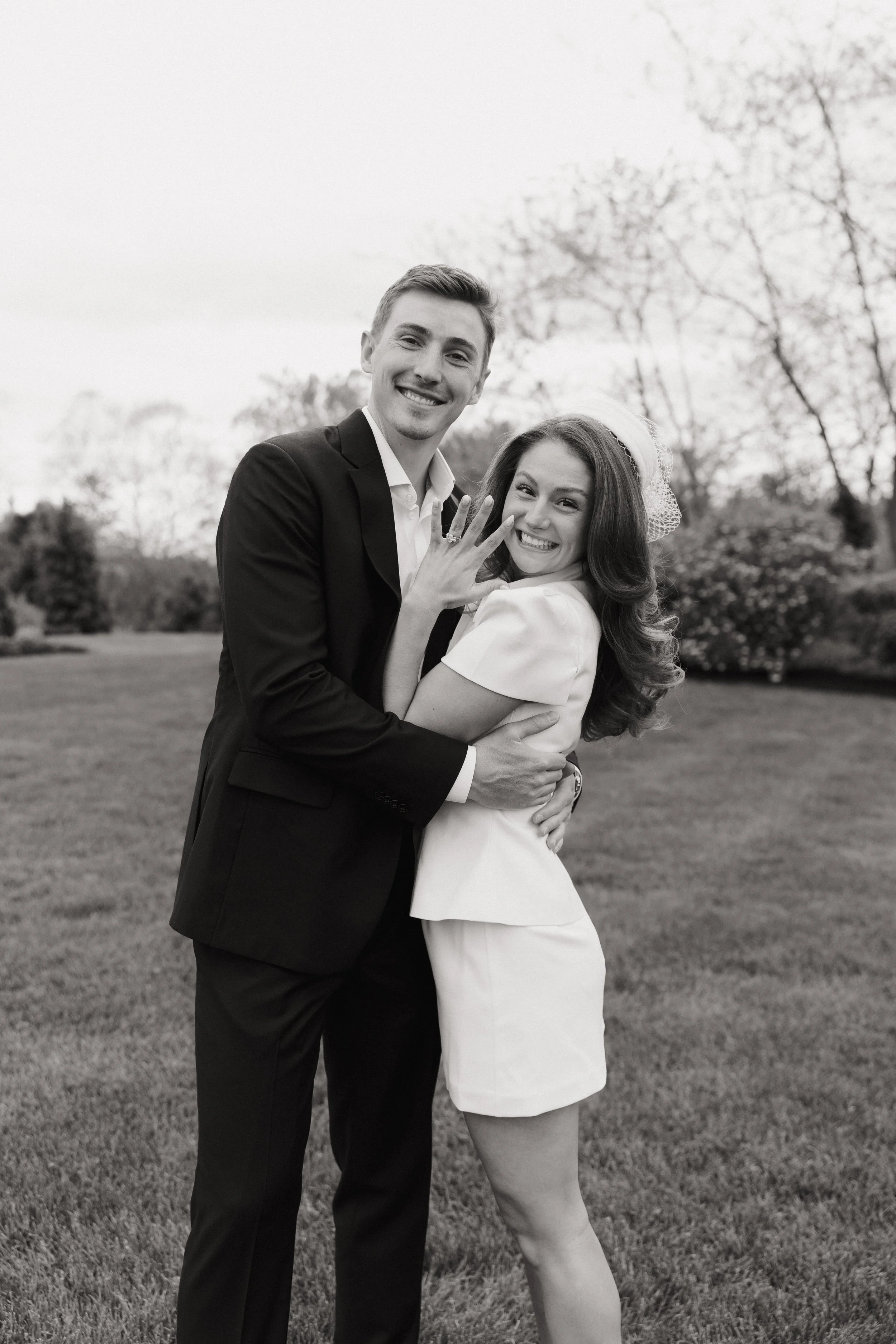 A smiling man and woman embracing outdoors with trees and grass in the background, woman displays engagement ring.