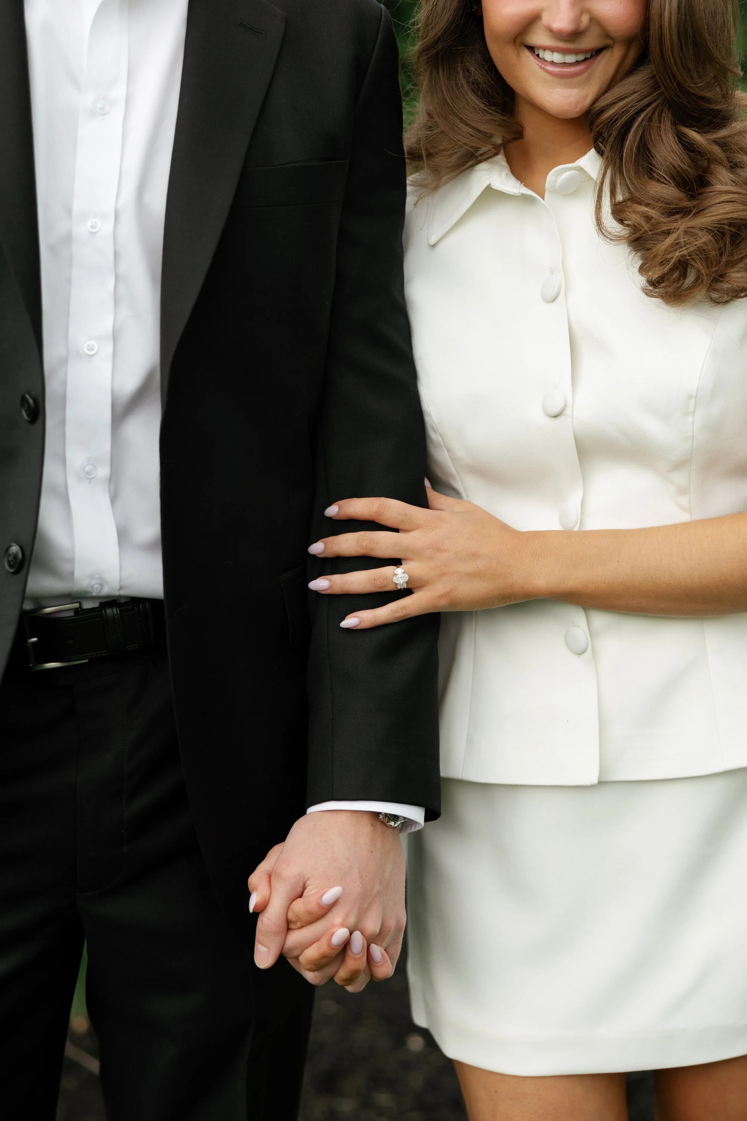 A close-up of a couple holding hands, with the woman wearing a white dress and a ring on her finger, and the man dressed in a black suit and white shirt, outdoors with greenery in the background.