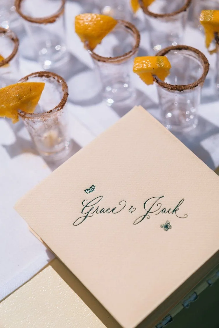 Wedding table setting with shot glasses garnished with lemon wedges, and a place card reading 'Grace & Jack' decorated with small butterfly illustrations.