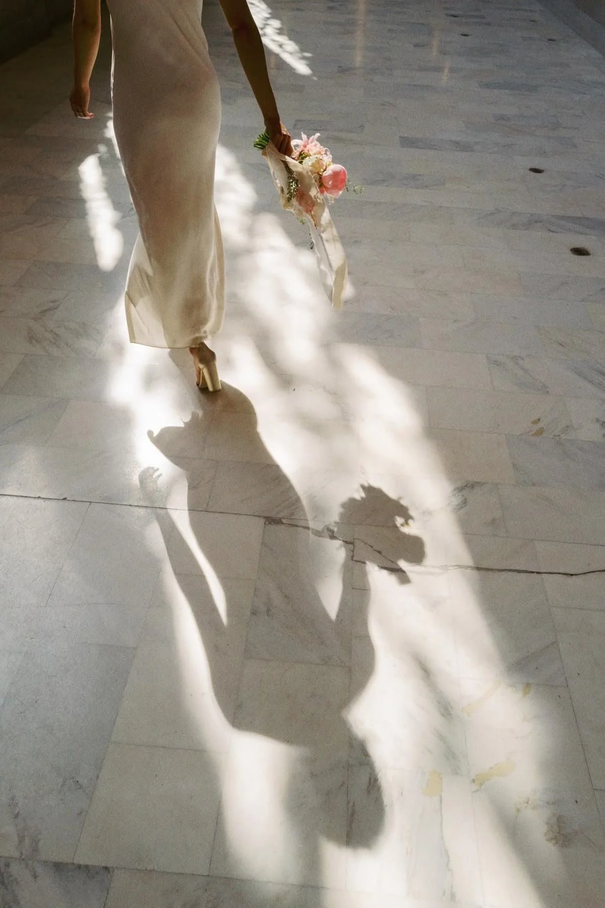 A woman in a beige dress holding a pink bouquet walking on a marble floor with sunlight creating shadows.