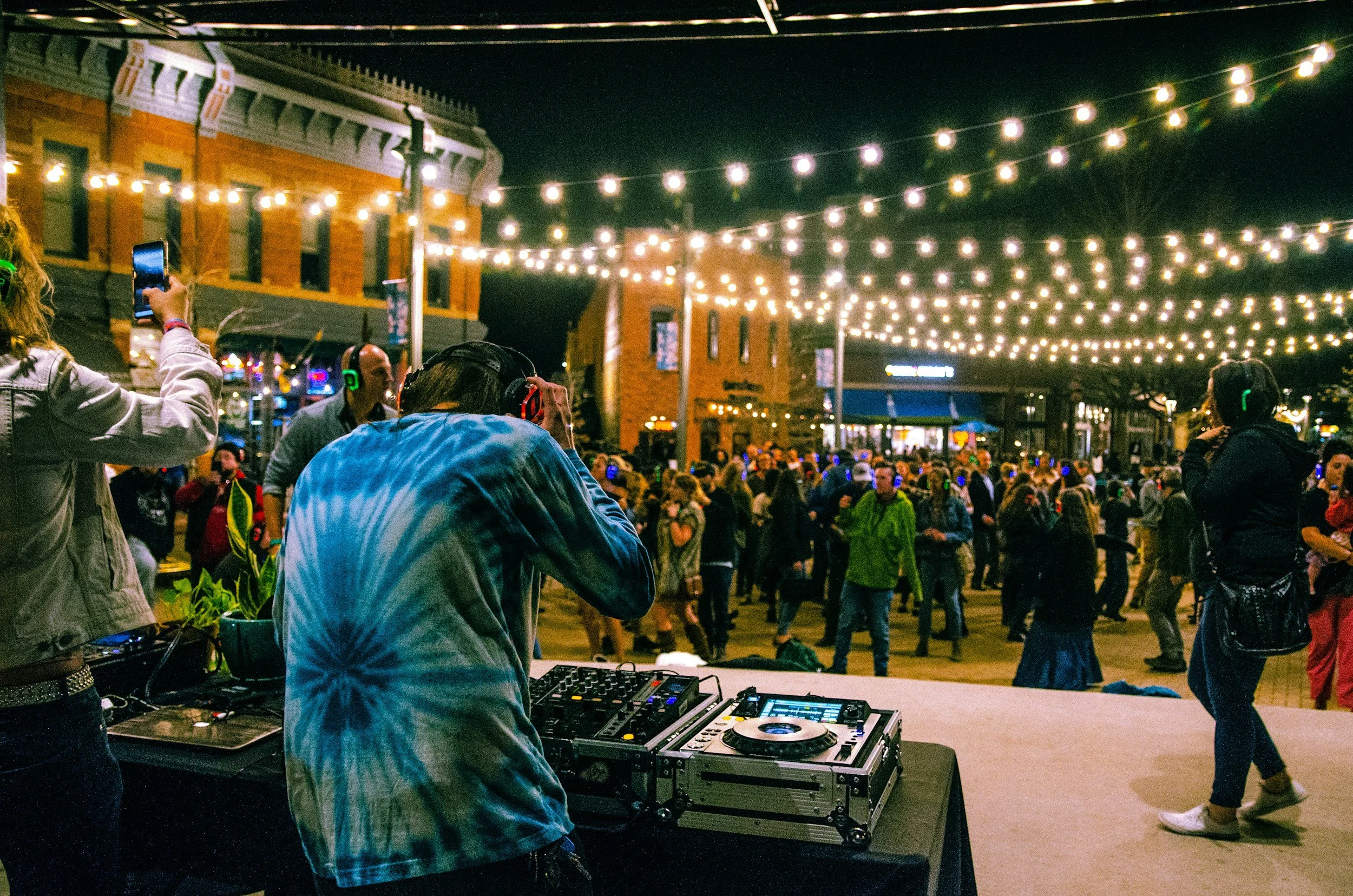 A DJ wearing a blue tie-dye shirt performs at an outdoor night event with string lights overhead. Multiple people, some with headphones, are dancing and taking photos or videos, with a brick building in the background.