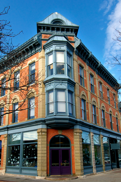 A historic four-story brick building with decorative metal details and large windows, featuring a rounded bay window on the corner, and a prominent rooftop with a decorative metal crest, set against a partly cloudy blue sky.