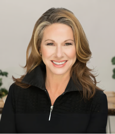 A smiling woman wearing a black zip-up top, indoors with a neutral background.