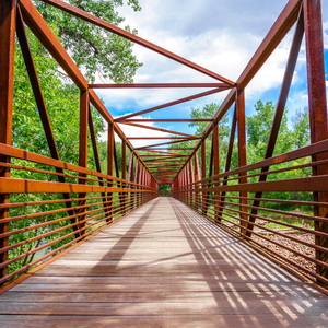 A wooden and metal footbridge with geometric overhead support beams, surrounded by green trees and a partly cloudy sky.