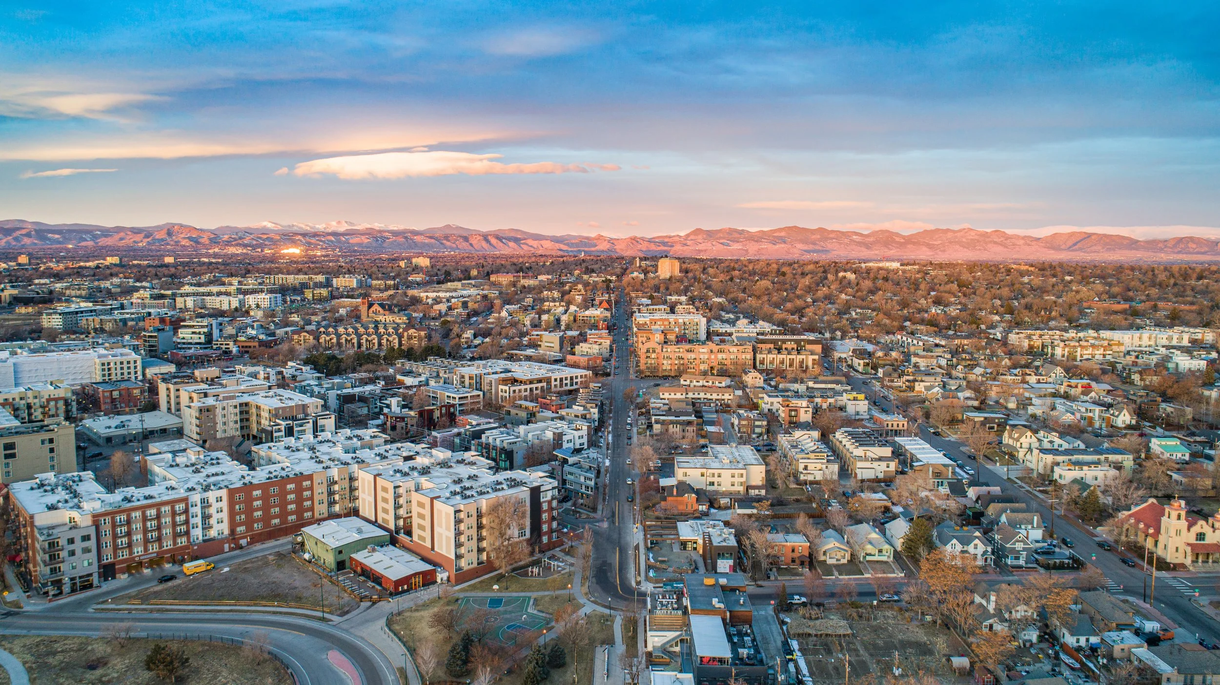 A cityscape at sunset with mountains in the background, showing a mix of residential and commercial buildings, roads, and trees.