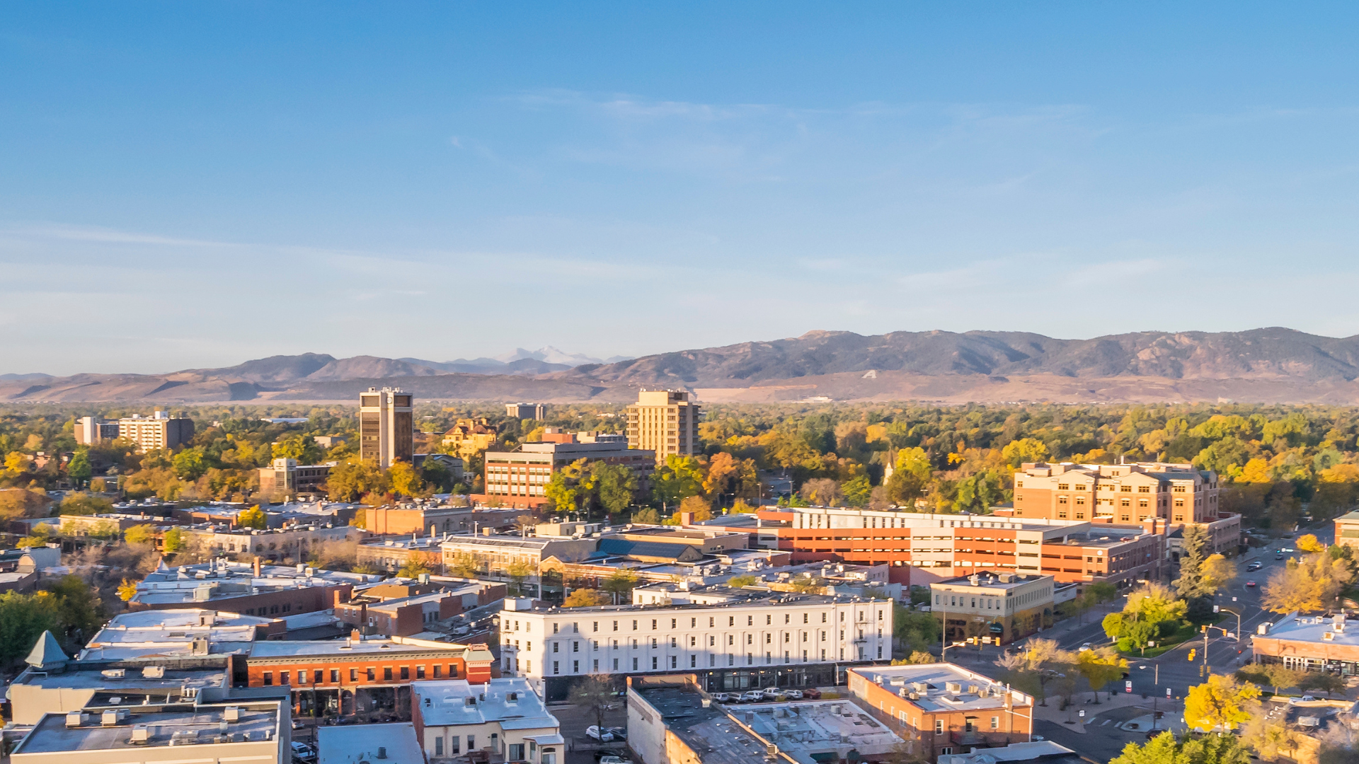 A cityscape with tall buildings, surrounded by trees with autumn foliage, and mountains in the background under a clear blue sky.
