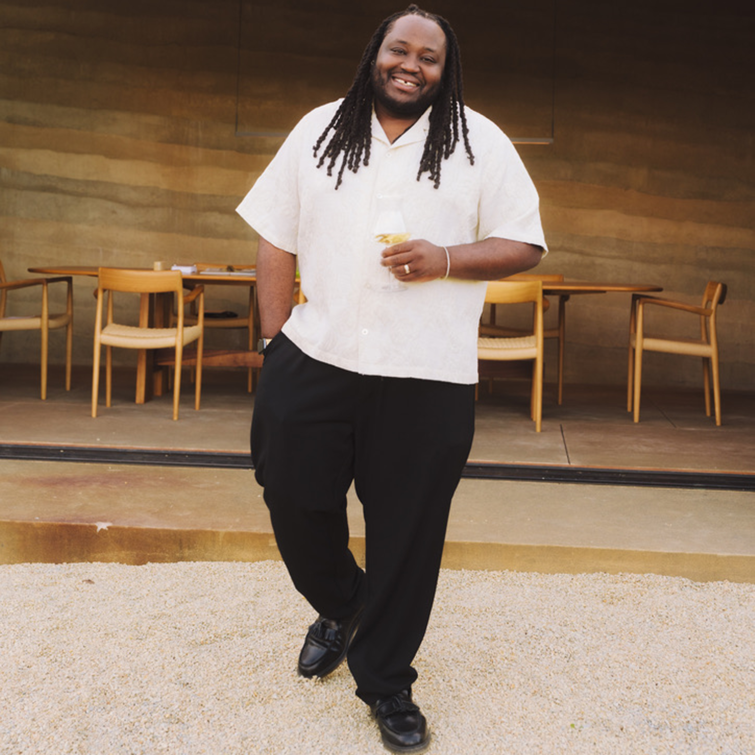 Smiling man with dreadlocks wearing a white short-sleeve shirt and black pants, holding a drink, standing outside a restaurant.
