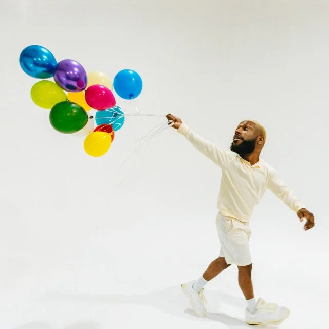 Man wearing a white shirt and shorts holding a bunch of colorful balloons in a white studio.