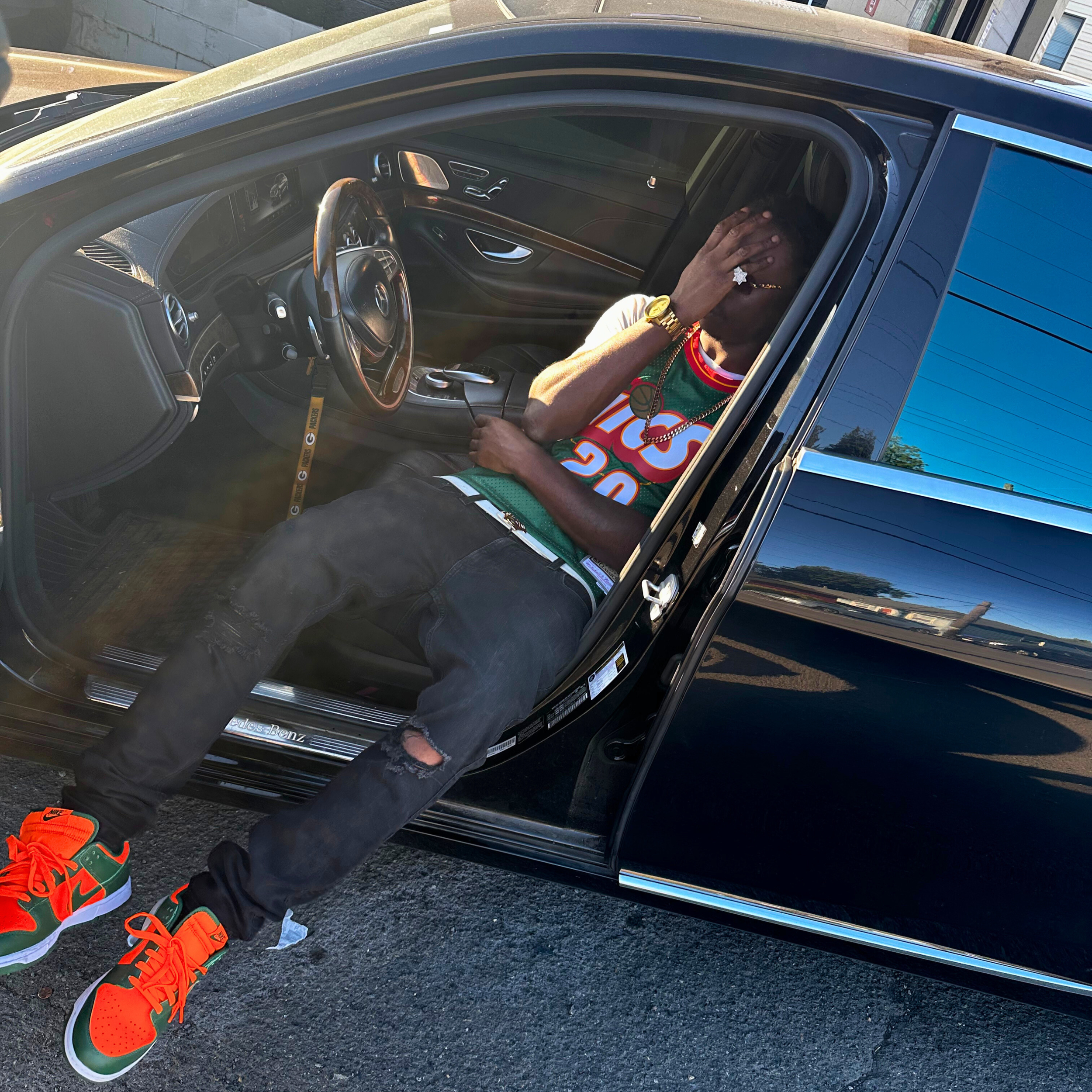 A young man sitting in the driver's seat of a luxurious black Mercedes-Benz car, wearing a colorful basketball jersey, ripped black jeans, and bright orange and green sneakers. He is accessorized with jewelry and a watch, and has his right hand covering his face.
