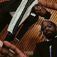 Two men in formal attire sitting in a room with wooden paneling, one wearing sunglasses and the other with glasses, with a hand in the foreground.