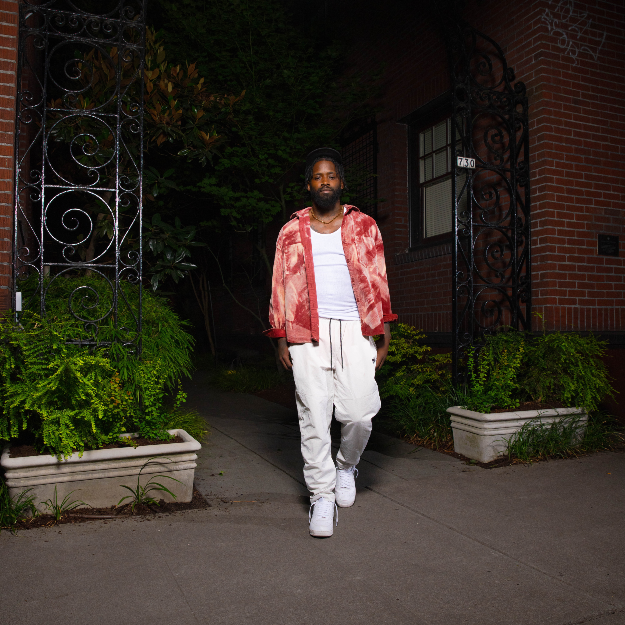 A man wearing a red patterned jacket, white shirt, and white pants walking on a sidewalk at night with greenery and a brick building in the background.