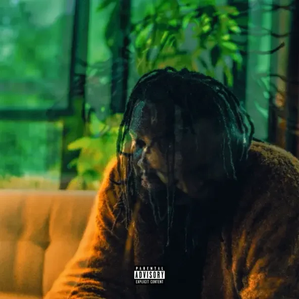 A person with dreadlocks sitting on a couch, looking thoughtfully to the side, with green plants and window in the background.