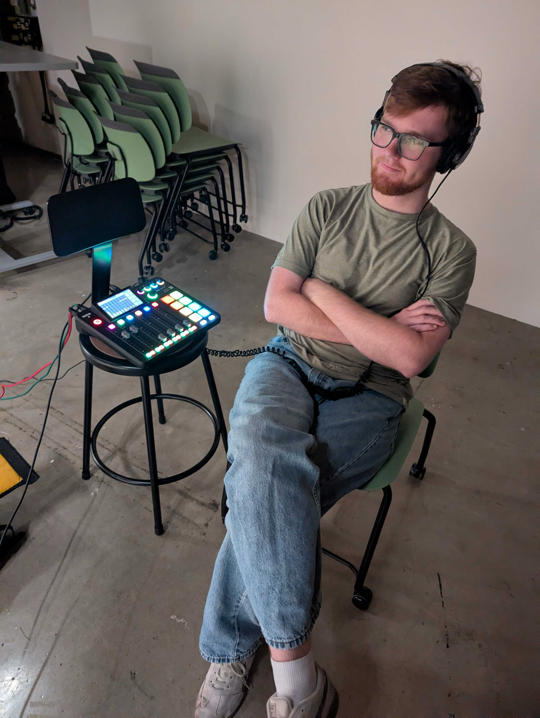 A young man with glasses, a beard, and headphones sitting in a chair with arms crossed, next to a table with a music production controller, in a room with stacked green chairs in the background.