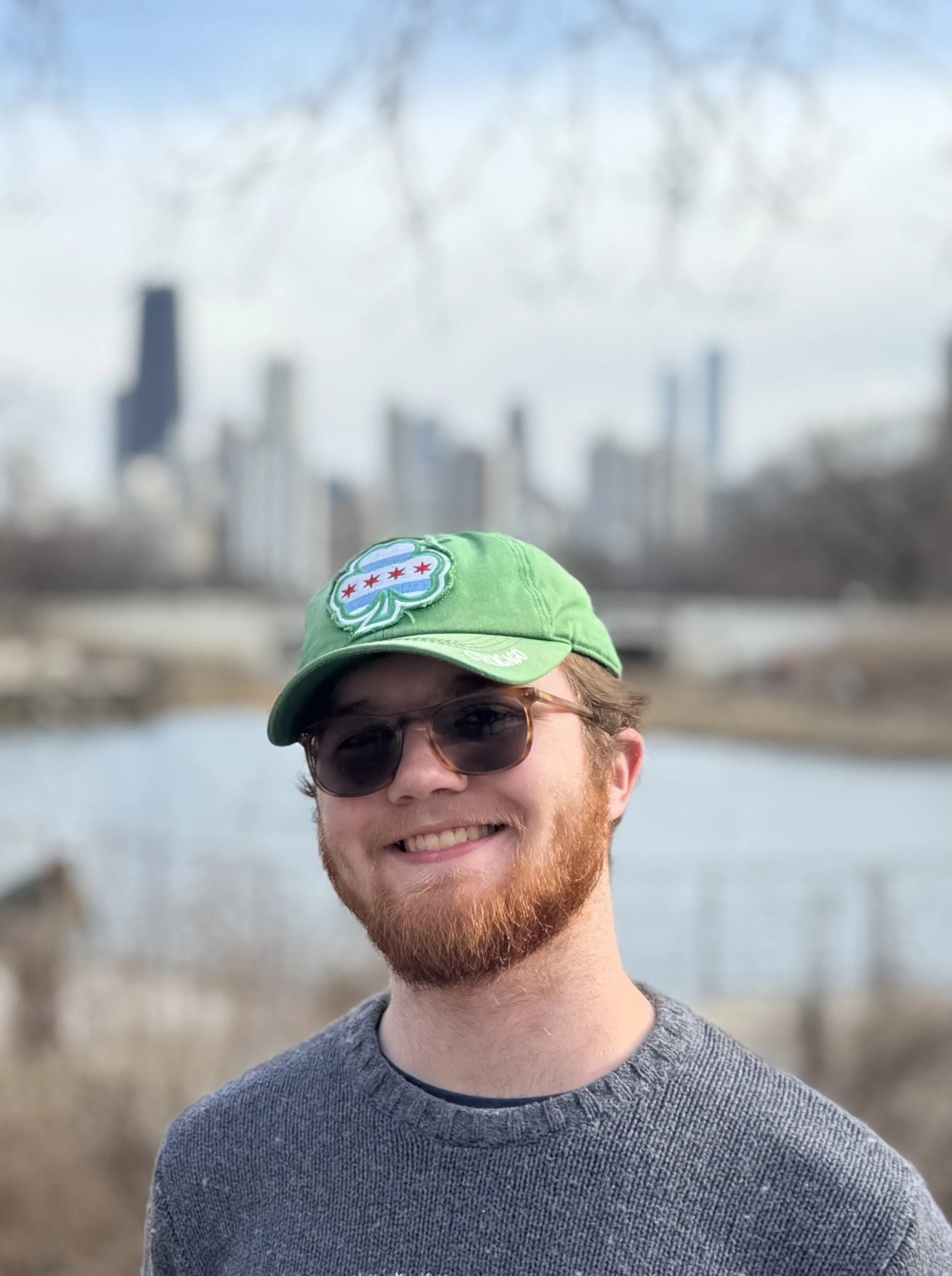 A smiling young man with a beard wearing sunglasses and a gray sweater, outdoors near a body of water with a city skyline in the background, including high-rise buildings and a smokestack, on a partly cloudy day.